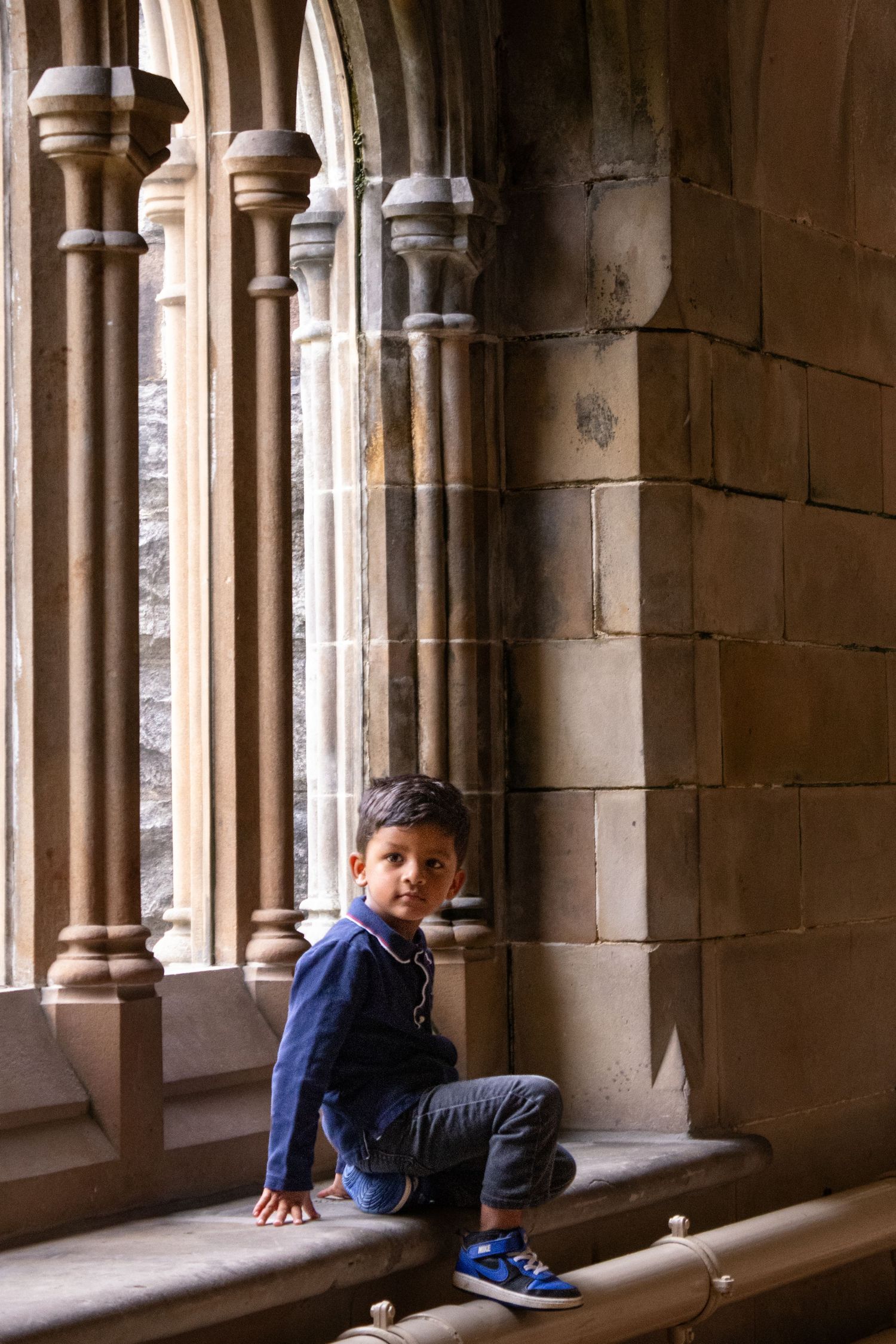 Young boy sitting on stone ledge in historic Gothic archway corridor with ornate columns.