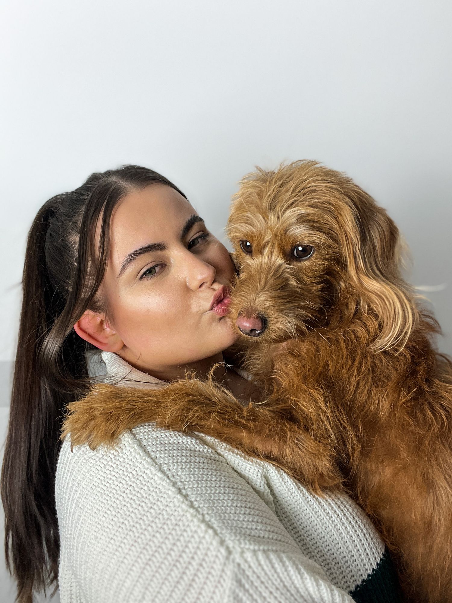 A fluffy brown dog receives a loving kiss while being held in a cozy white sweater.