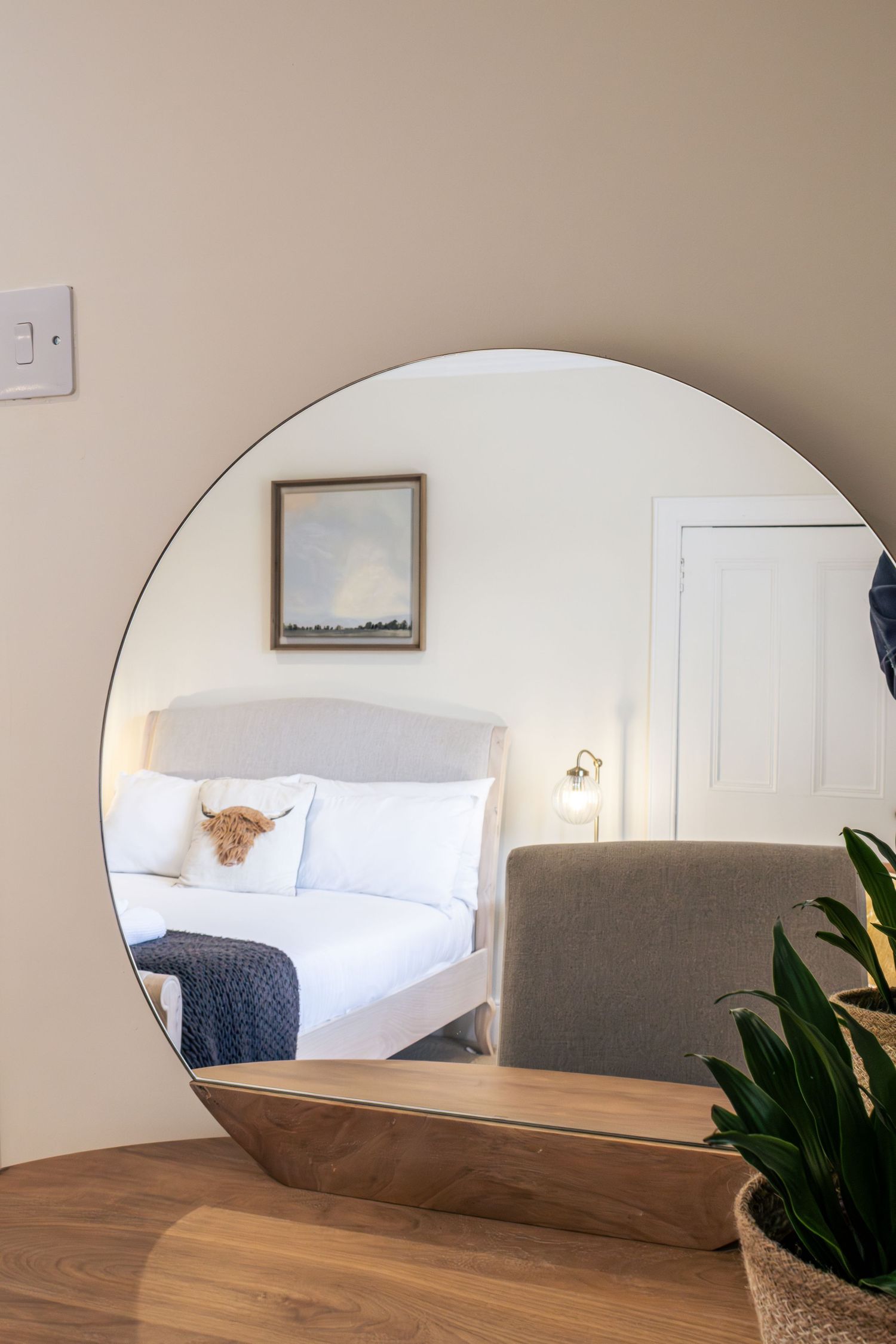 Round mirror reflecting a cozy bedroom with white bedding, highland cow pillow, framed art, and wall sconce lighting.