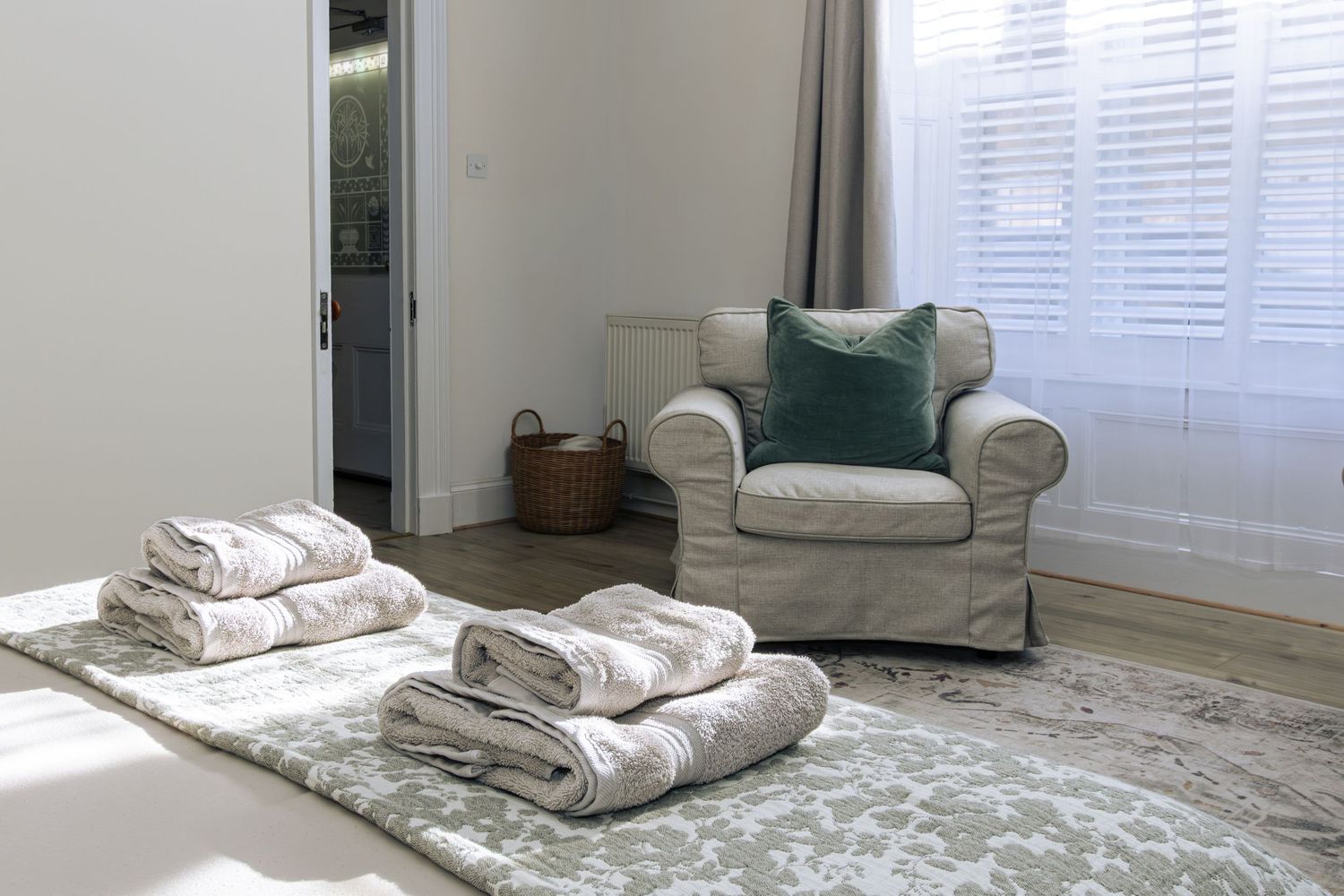 Bright bedroom with a white armchair, green cushion, folded towels on a patterned rug, and natural light from shuttered windows.