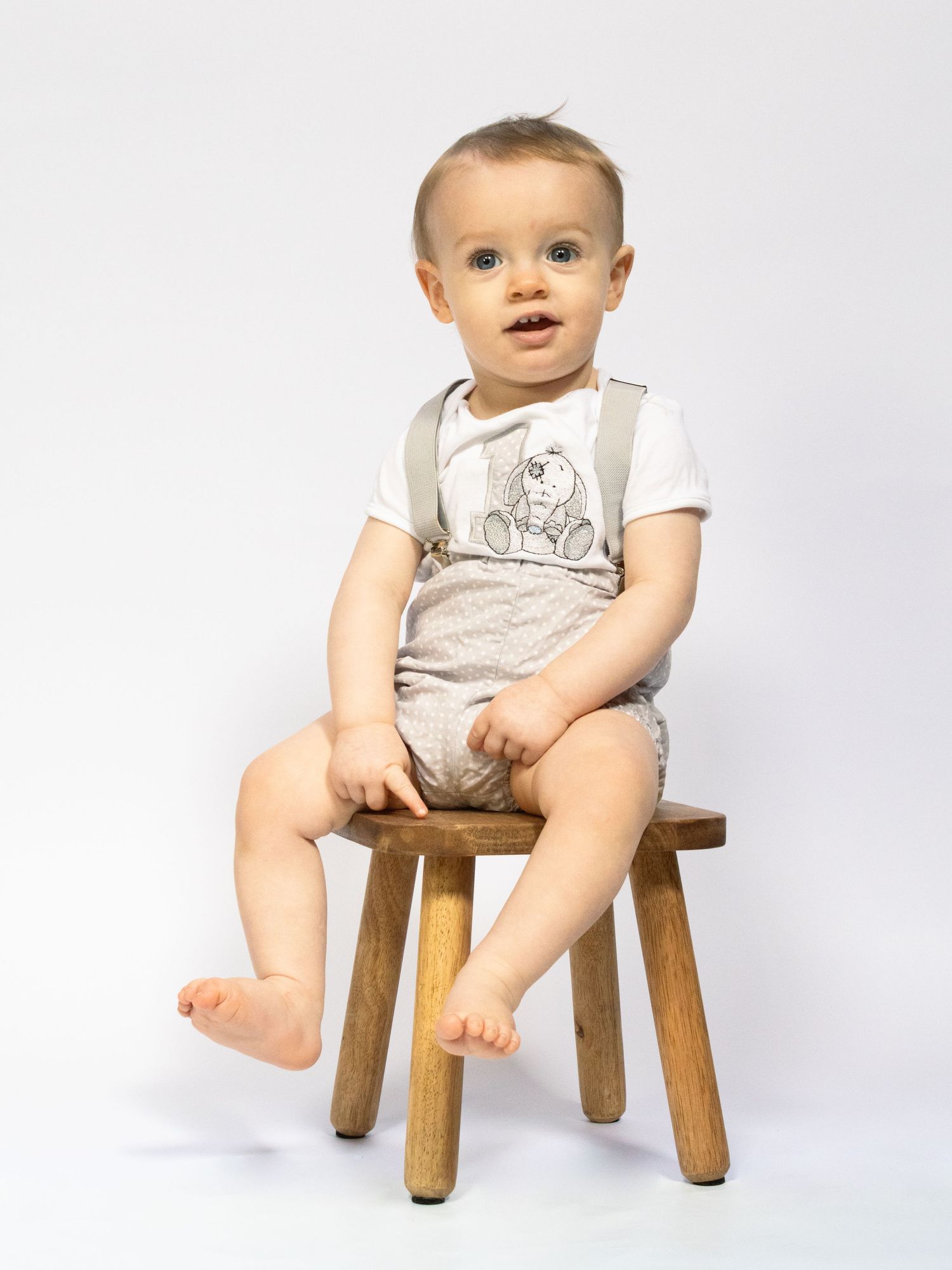 Cute baby boy sitting on a wooden stool wearing suspenders and a teddy bear graphic outfit.