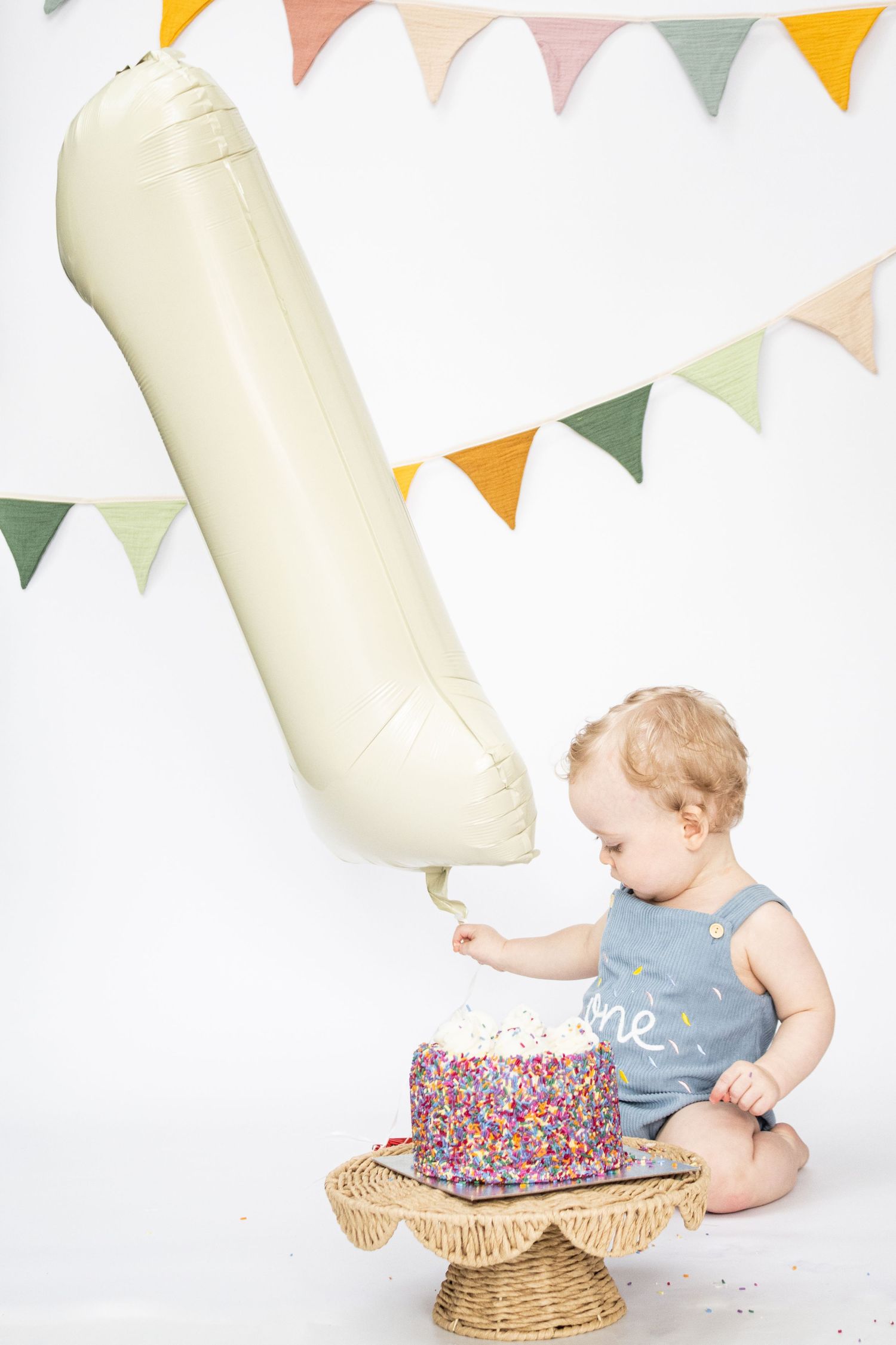 Baby in blue 'one' romper touching colorful sprinkle birthday cake beside number 1 balloon with festive bunting backdrop.