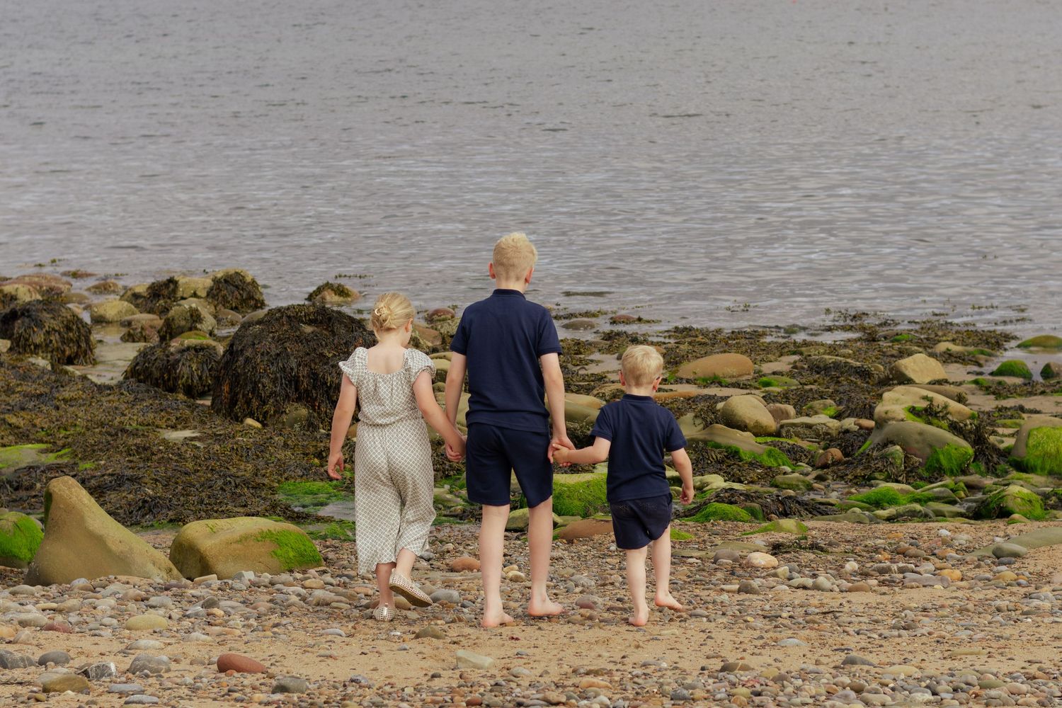 Three children holding hands on a rocky beach shoreline with green moss-covered stones and calm sea water.