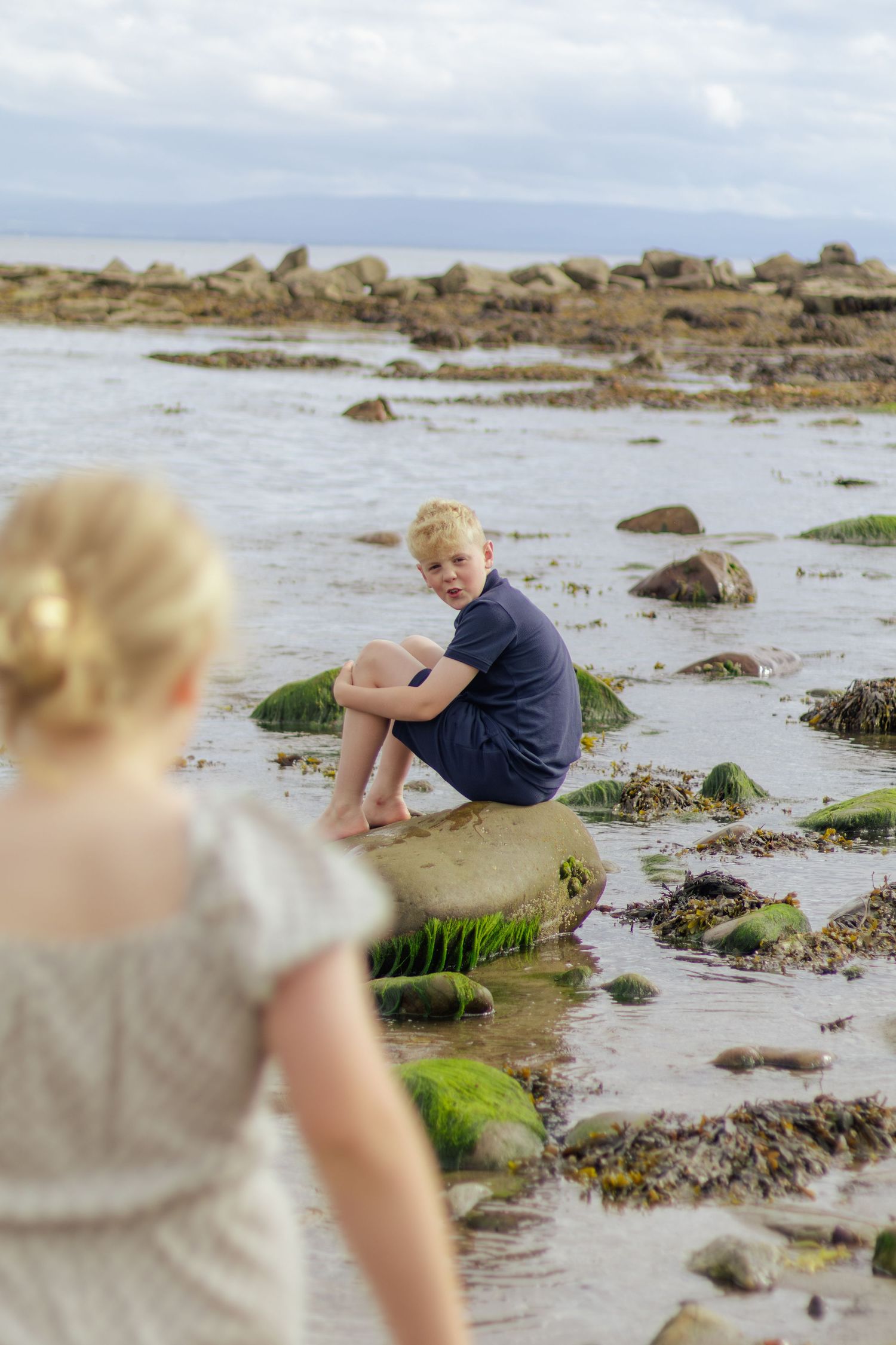A blonde boy sits on a mossy rock at a rocky shoreline while a girl in a white dress watches him from the foreground.