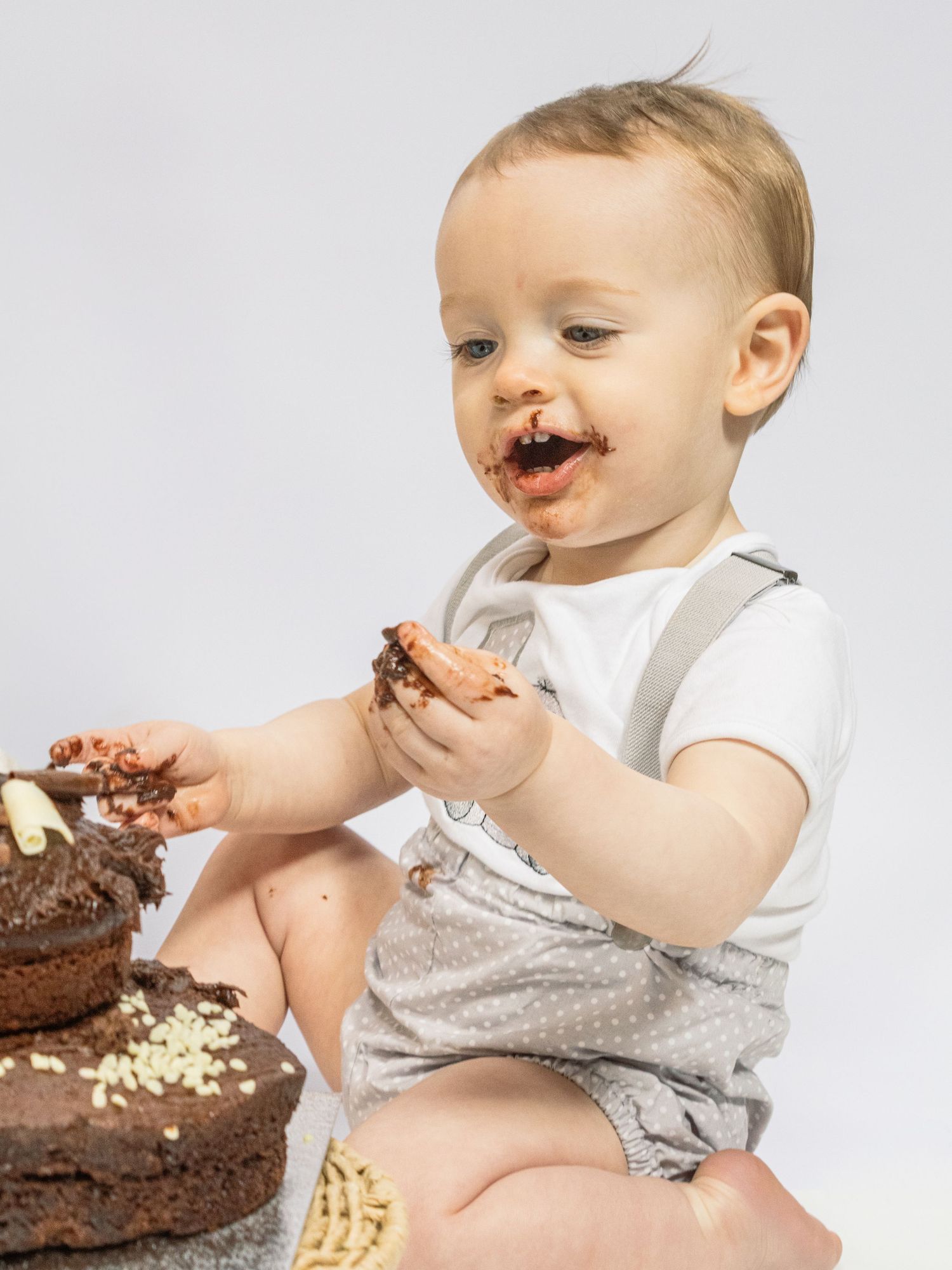 Baby with chocolate cake smash, smiling with frosting on face, wearing gray suspender romper.
