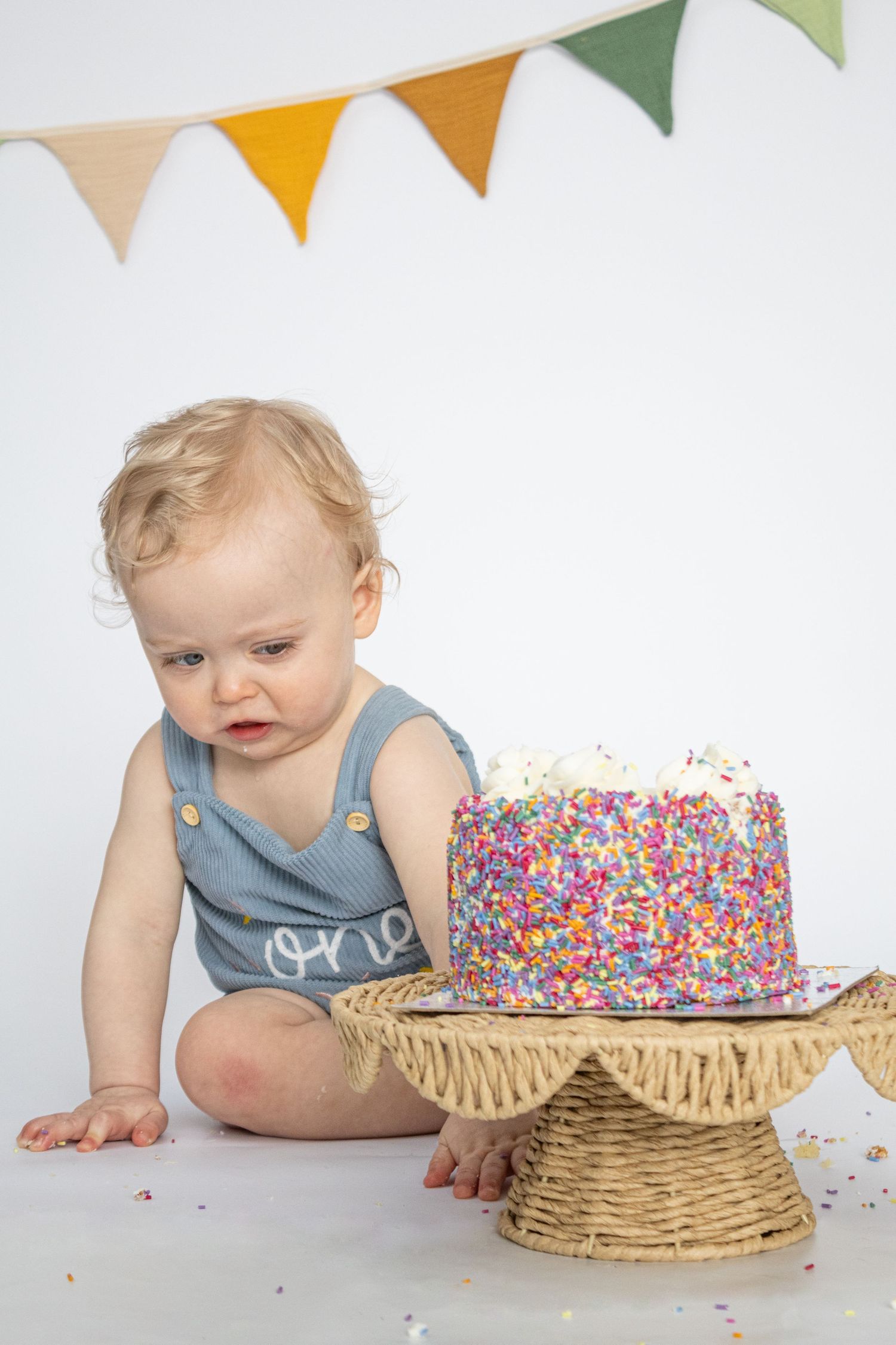 Baby in a blue “one” romper looking at a colorful sprinkle cake on a wicker stand.