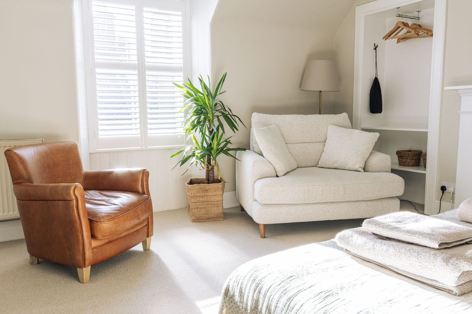 Bright bedroom sitting area with tan leather armchair, cream sofa, indoor plant, and white shuttered window.