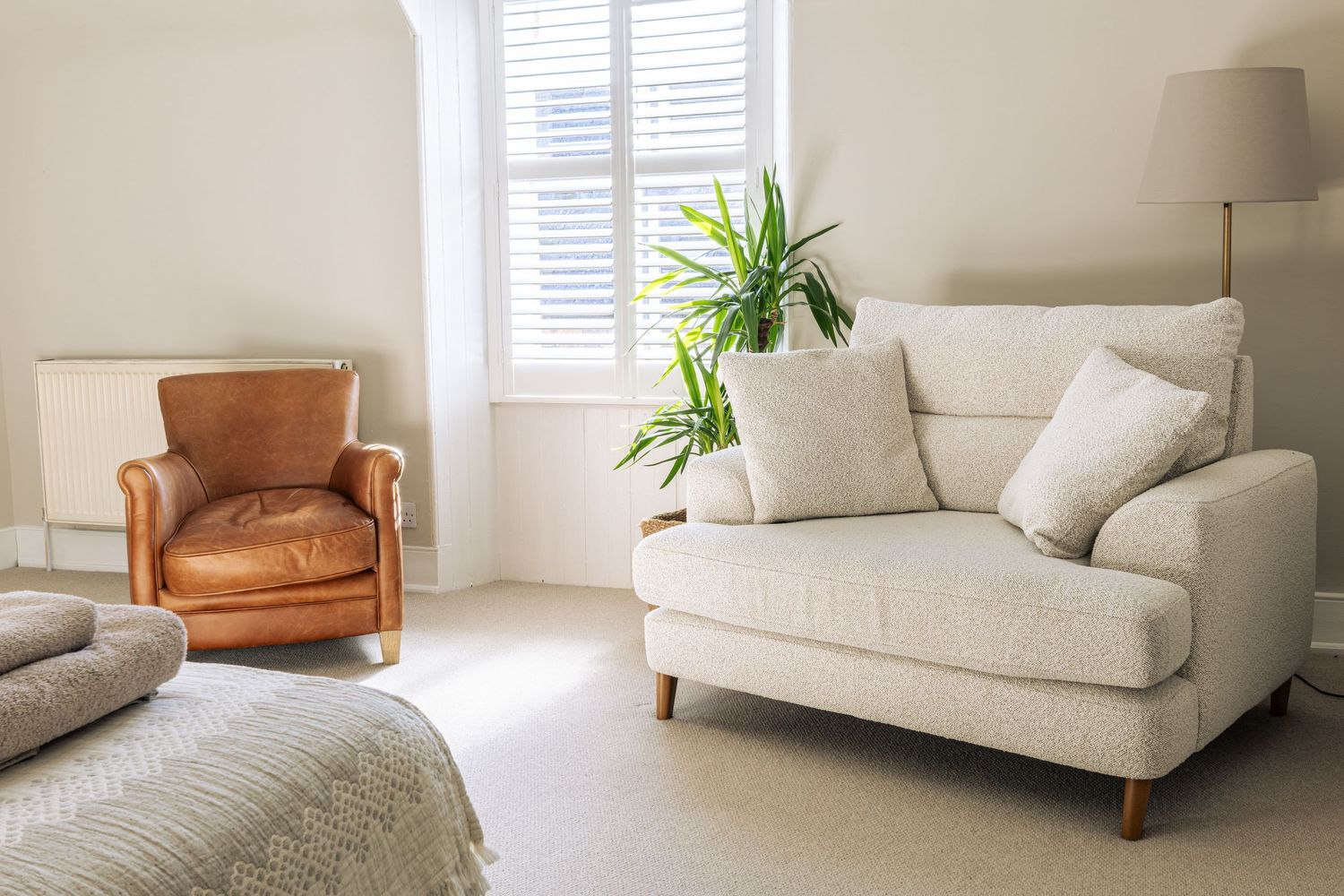Cozy living room with cream fabric loveseat, brown leather armchair, potted plant, and neutral carpet flooring near a window.