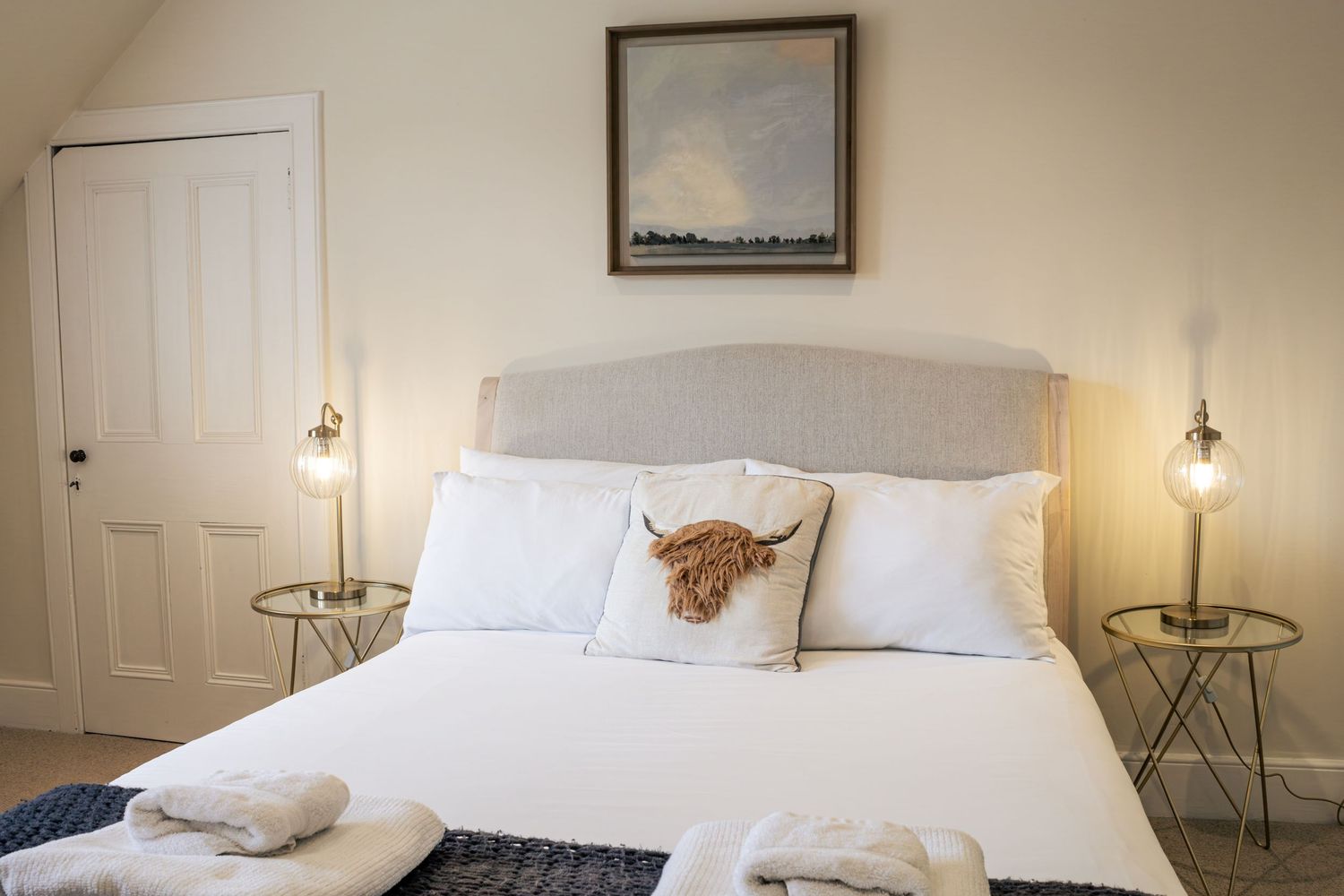 Cozy white bedroom with upholstered headboard, Highland cow pillow, flanking gold side tables with warm Edison bulb lamps.