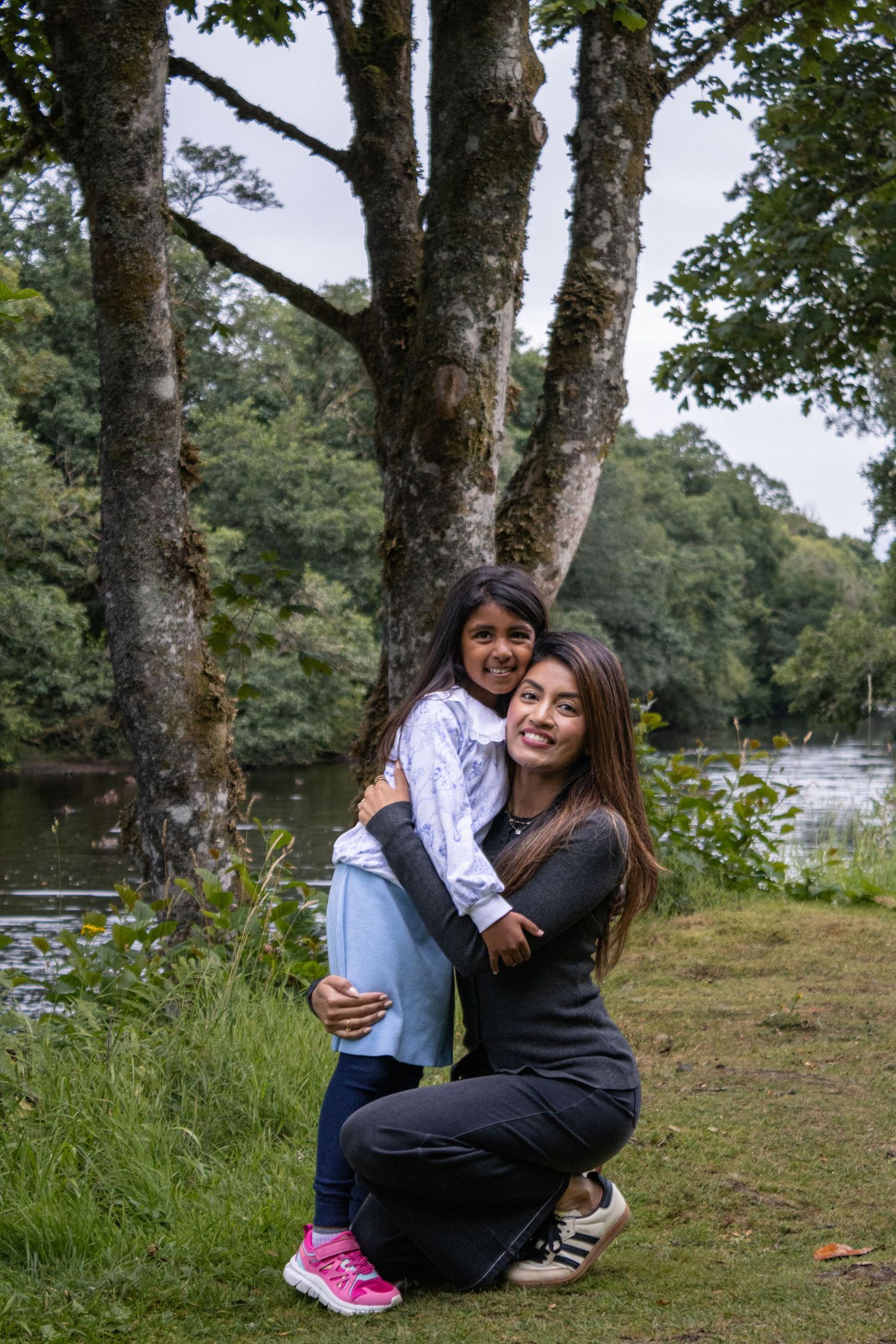 A young woman crouches hugging a smiling little girl near a river surrounded by lush green trees.
