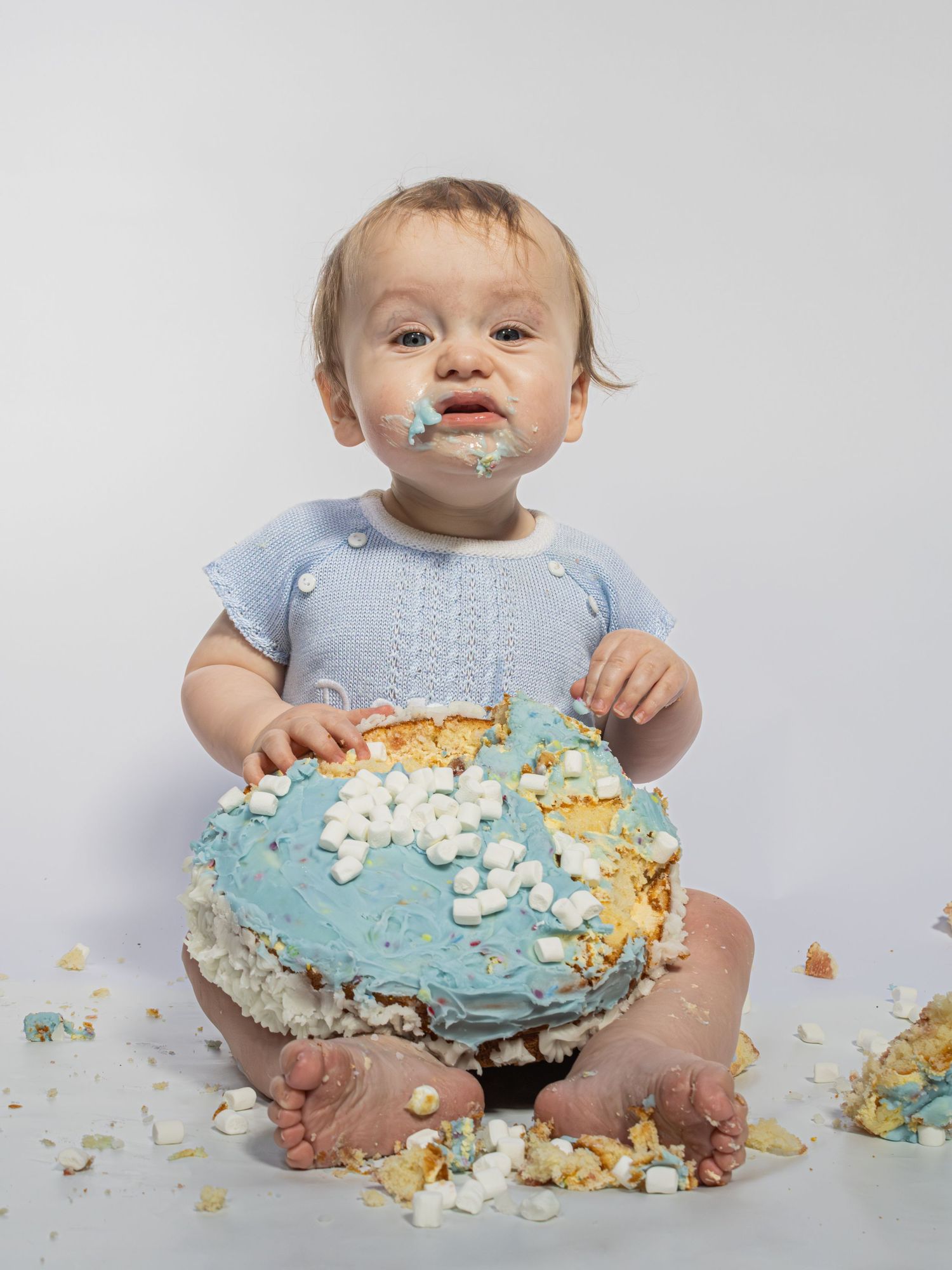 Baby in blue romper smashing a blue frosted first birthday cake, with frosting on face and cake crumbs scattered around.