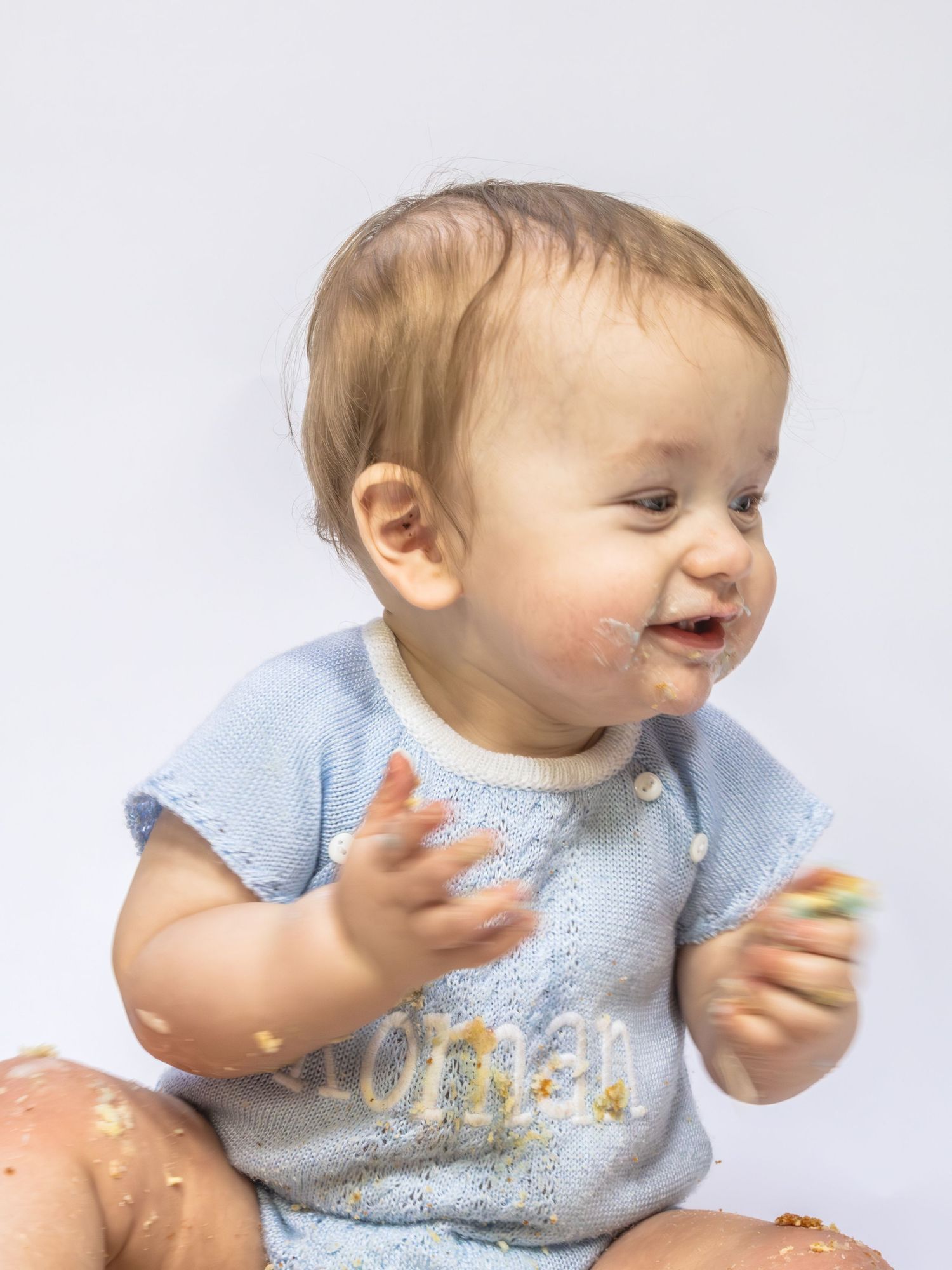 Happy baby boy covered in cake frosting clapping hands during first birthday cake smash celebration.