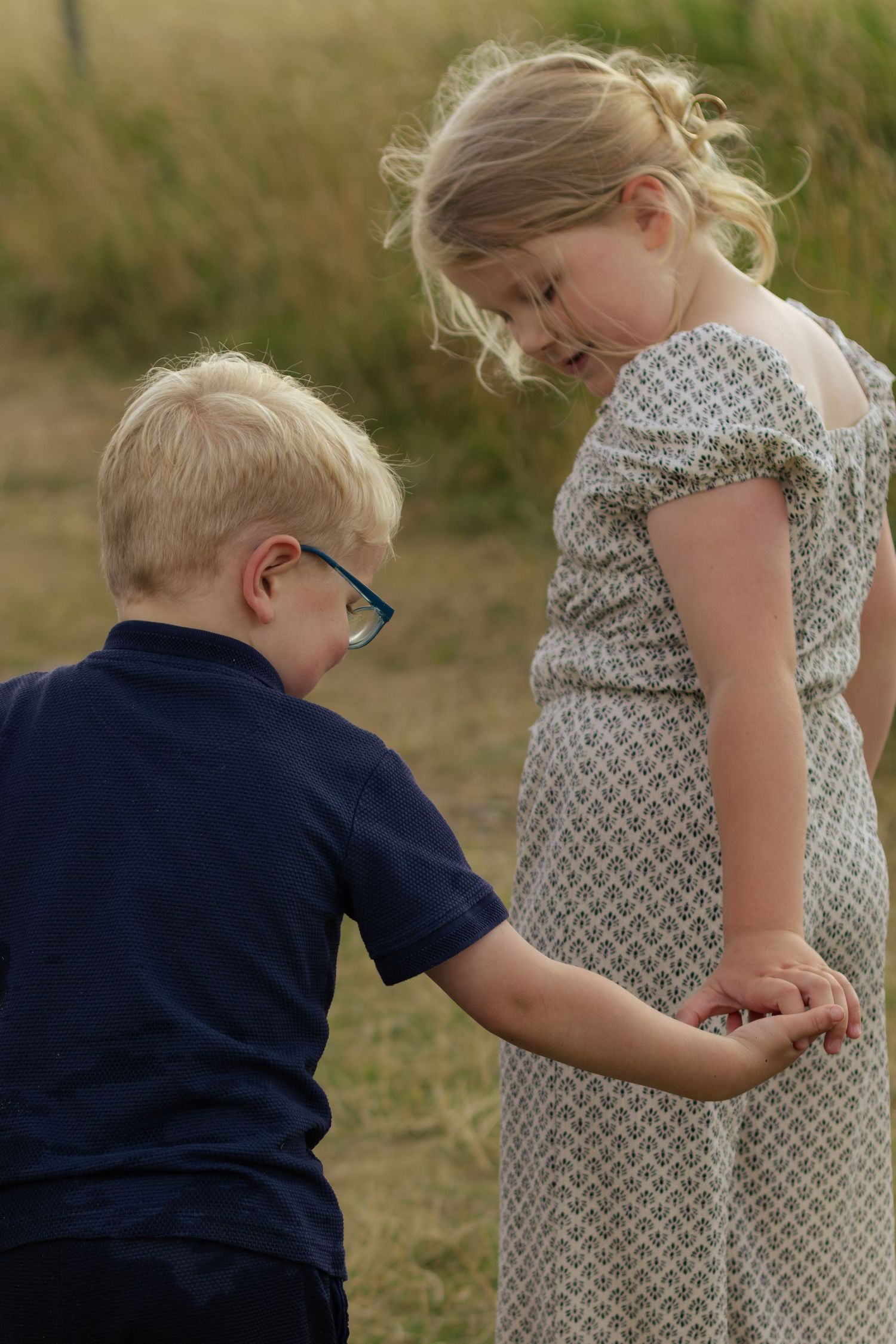 Two blonde children examining something in their hands outdoors in a field, boy wearing glasses and navy shirt.