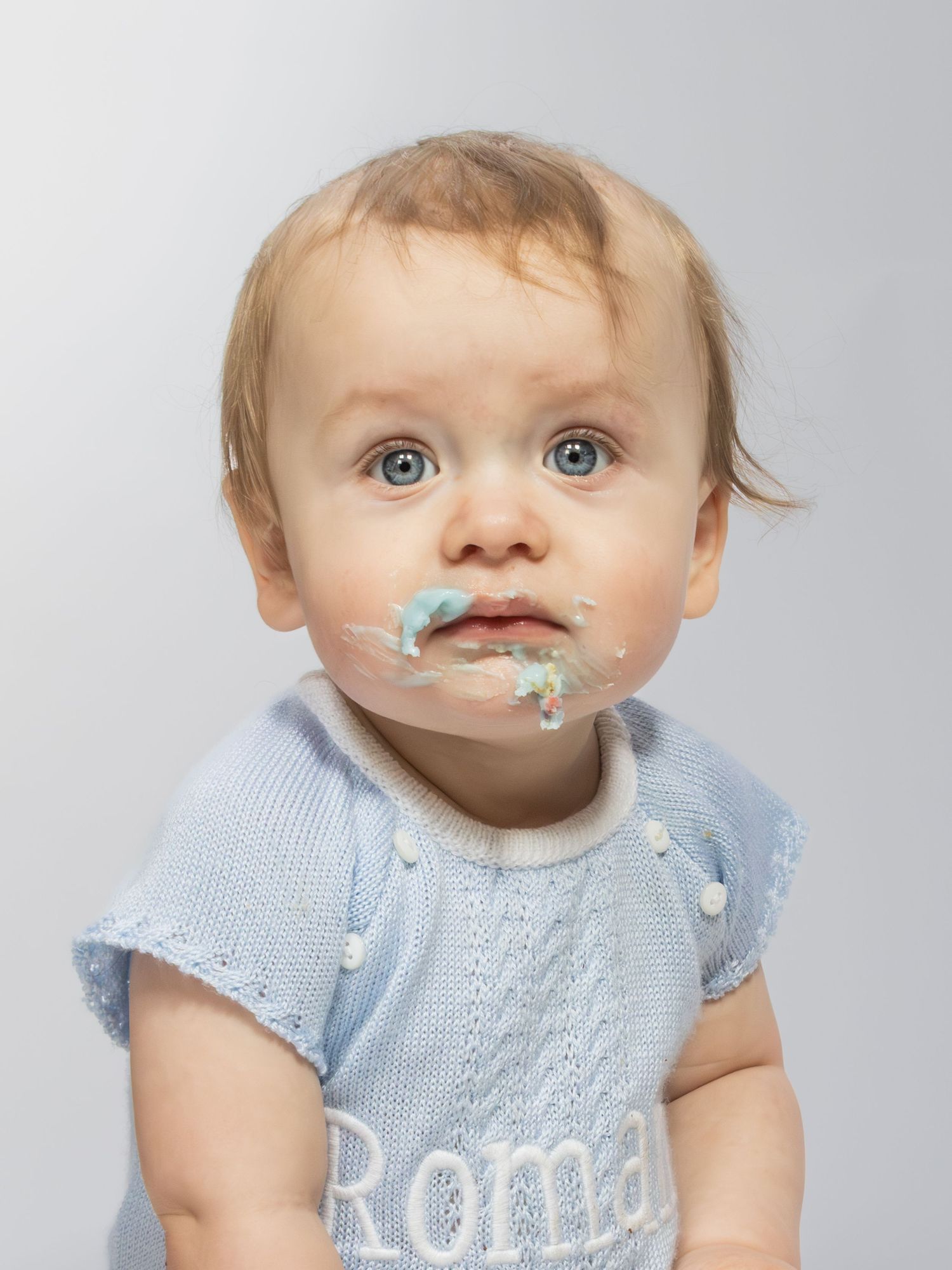 Cute baby named Roma with blue eyes and frosting on face wearing a light blue knit sweater against gray background.