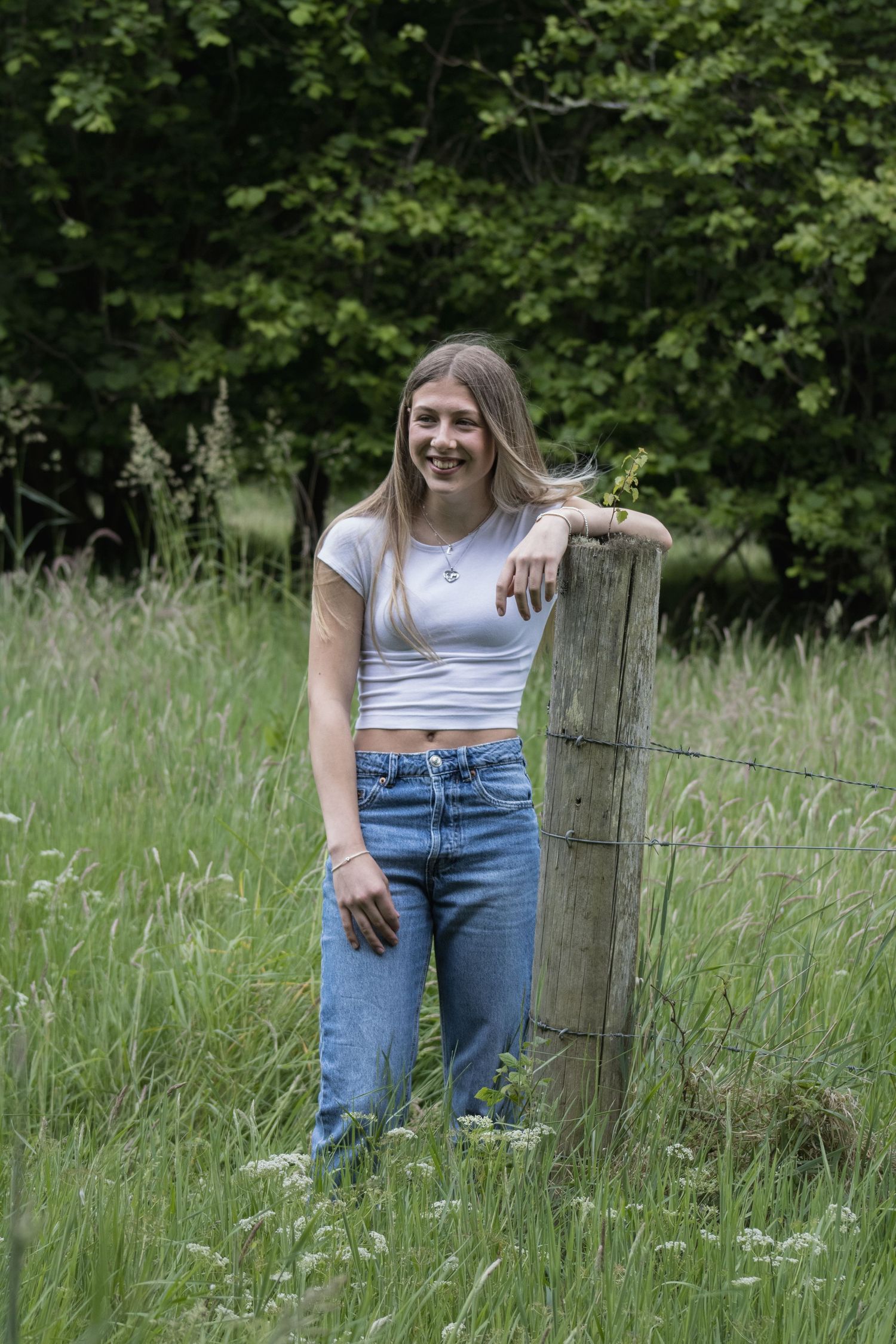 Person in casual attire poses playfully by wooden fence post in tall grass field.