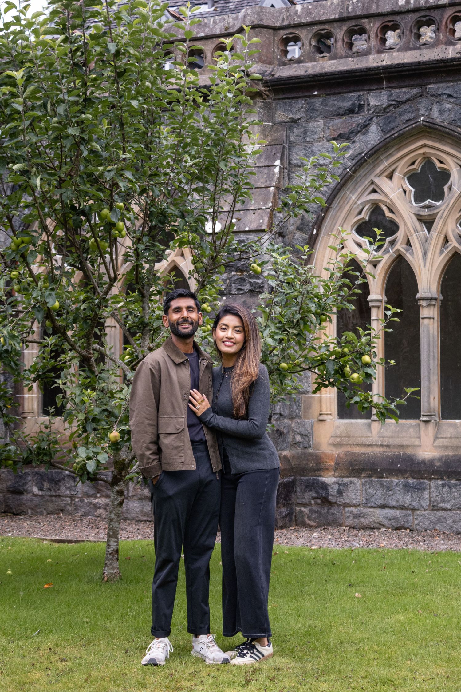 A smiling couple poses together on green grass before a Gothic stone building with apple trees.