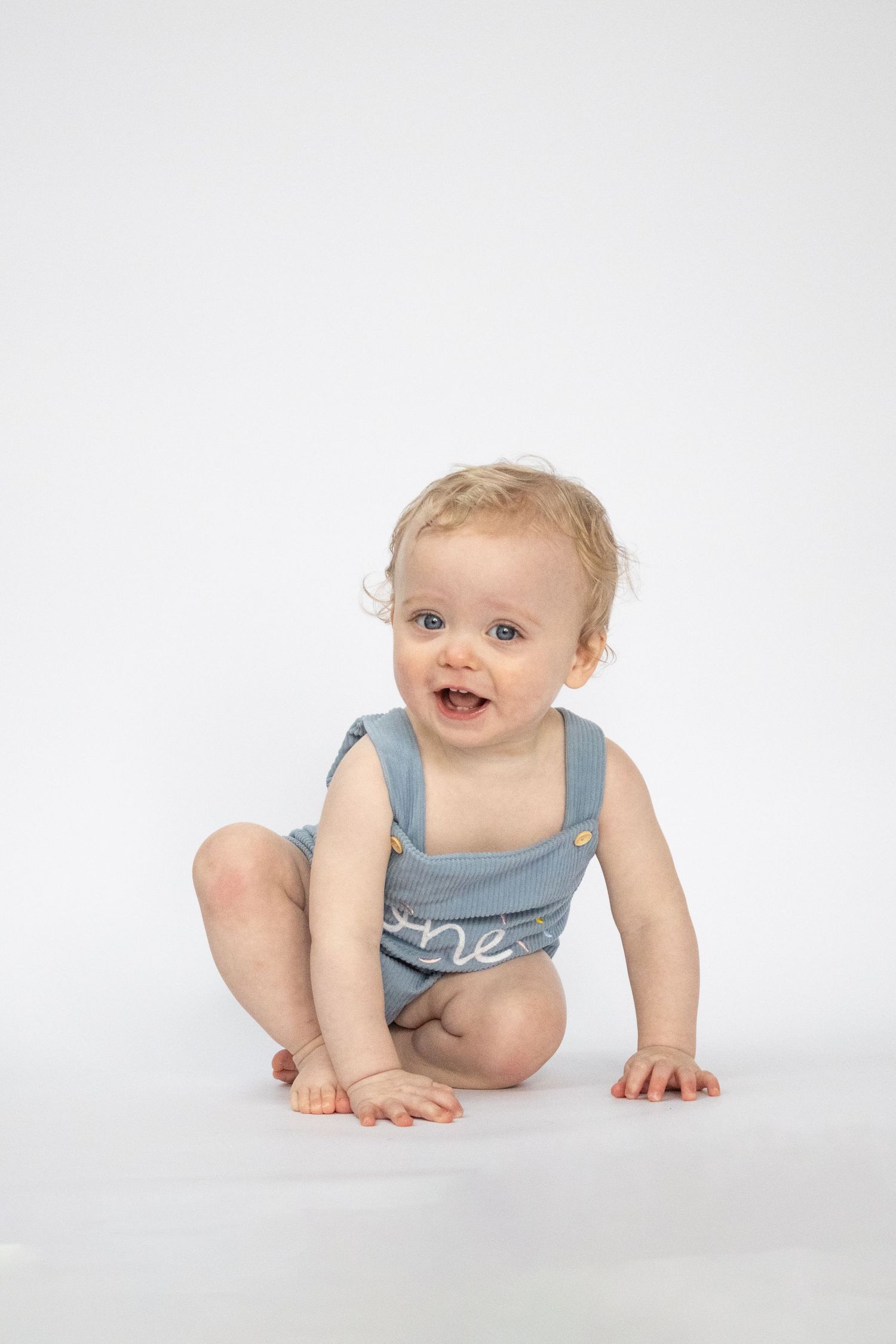 Smiling baby in a blue “one” romper sitting on a white background.