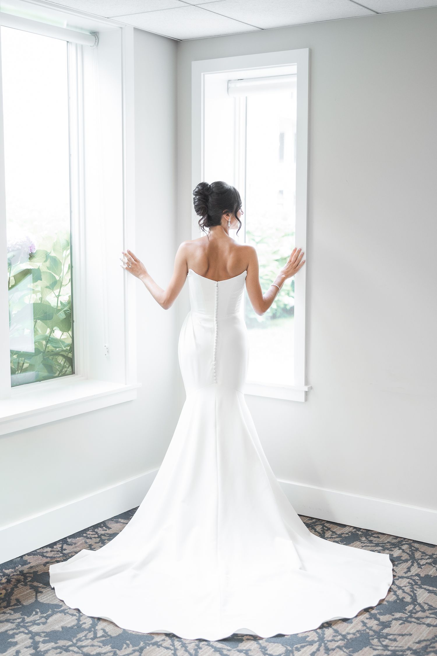 Bride in flowing white gown poses gracefully in bright room with multiple windows.