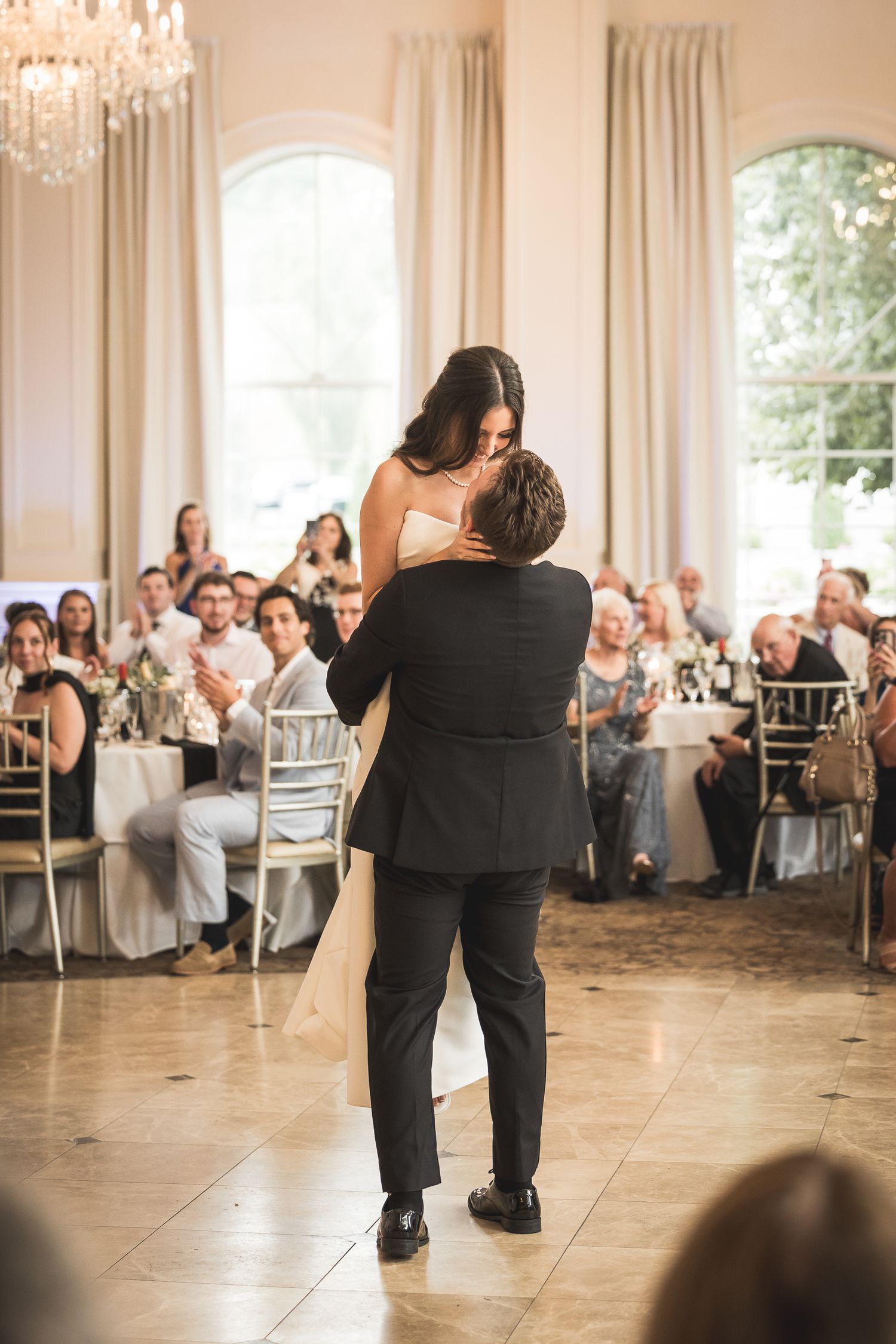 Wedding party dancing in an elegant ballroom with white curtains and chandeliers.