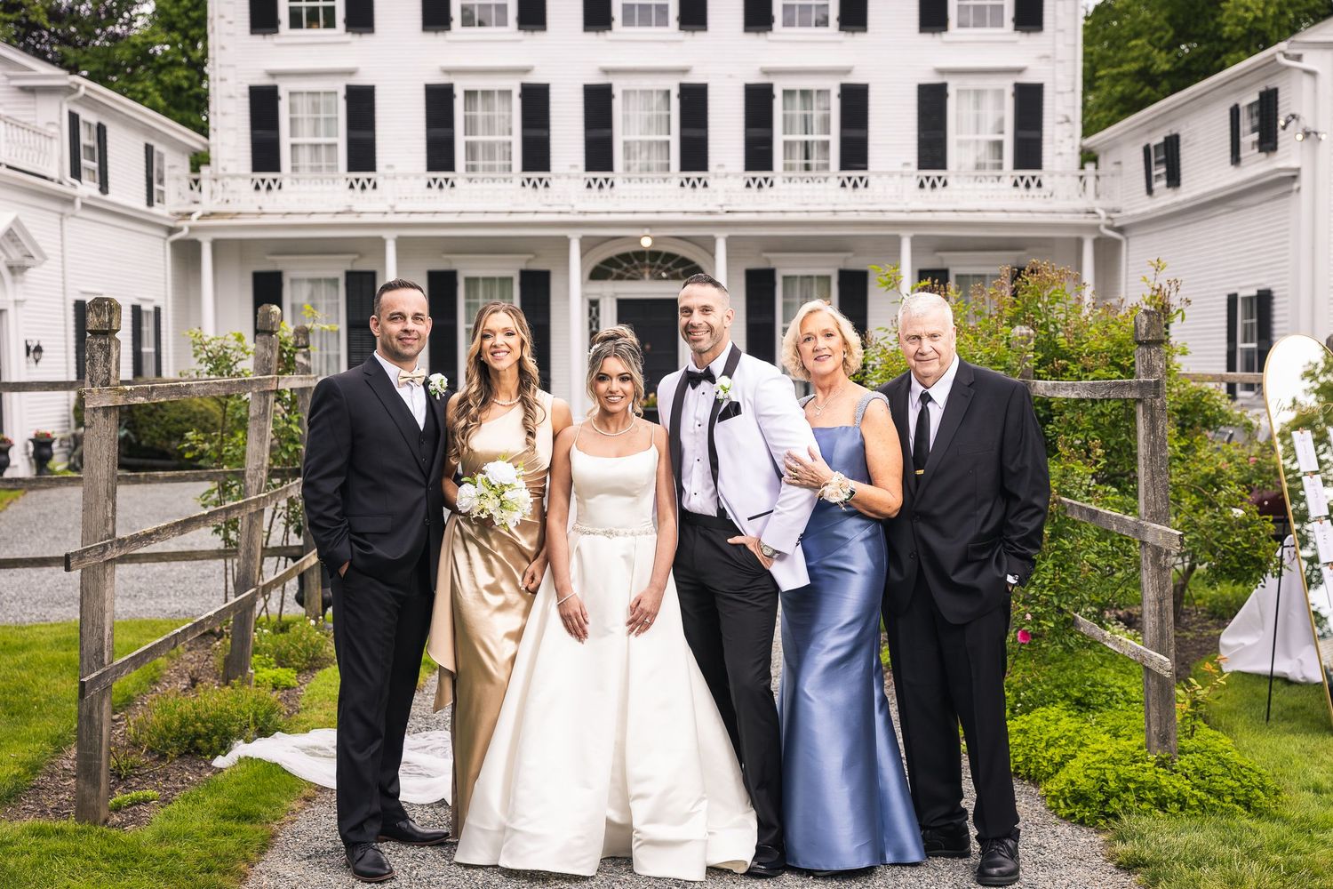 Wedding group poses along rustic fence with classic white colonial building in background.