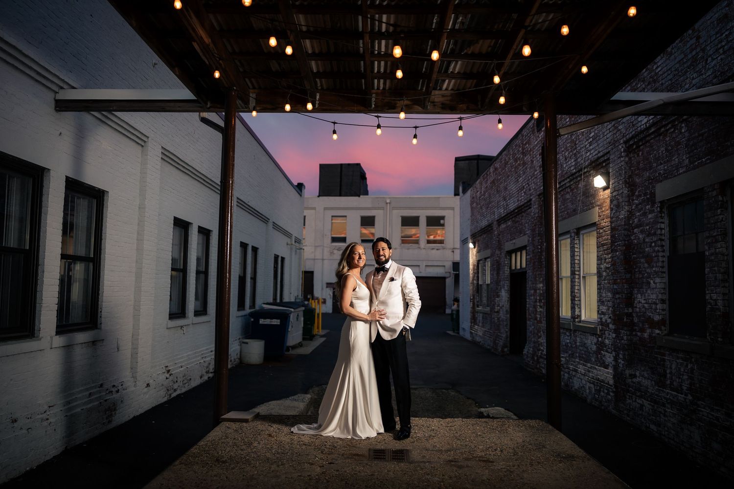 A wedding couple stands silhouetted in an industrial brick alleyway at dusk with warm lighting and dramatic shadows.