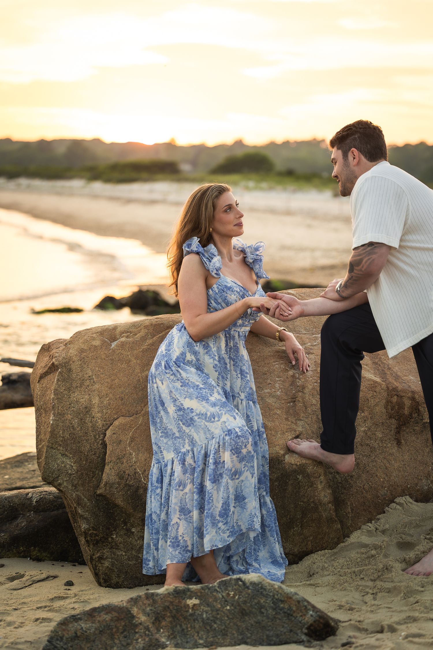 A couple shares a romantic moment on a rocky beach at sunset as they sit together near large boulders.