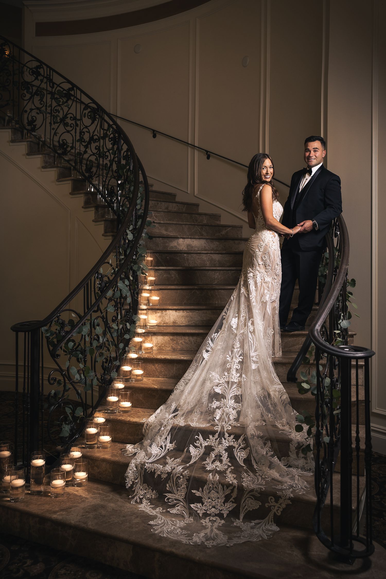 Romantic wedding couple poses on candlelit staircase with ornate iron railing and bride's lace train flowing behind.