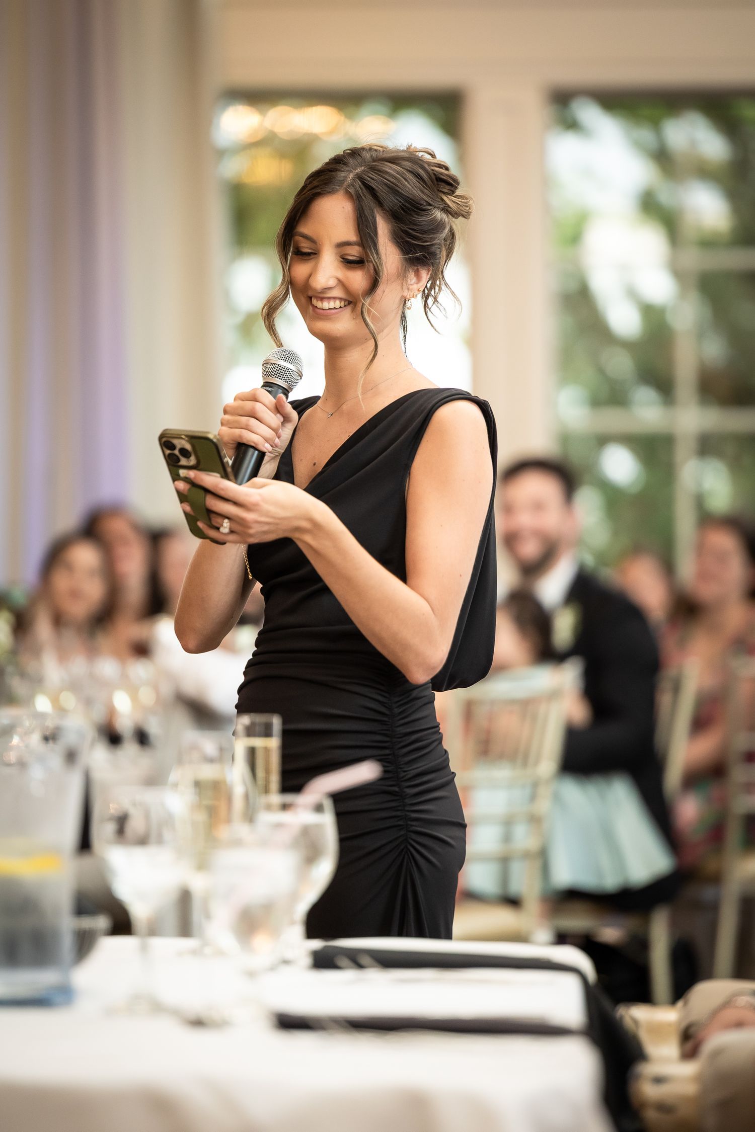 Wedding reception guest in black dress smiles while looking at phone during celebration.