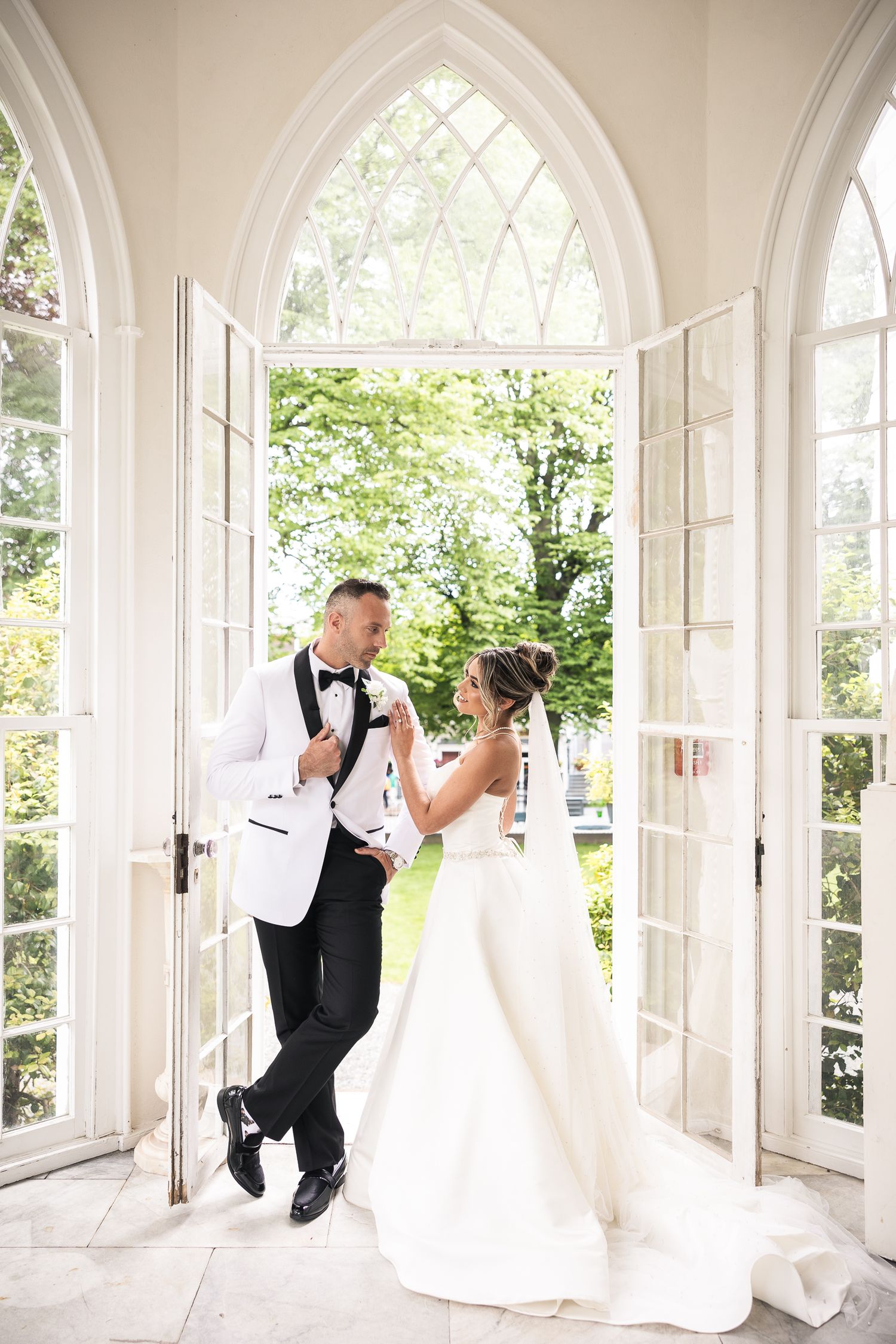 Romantic wedding couple poses in elegant white archway with natural light streaming through windows.