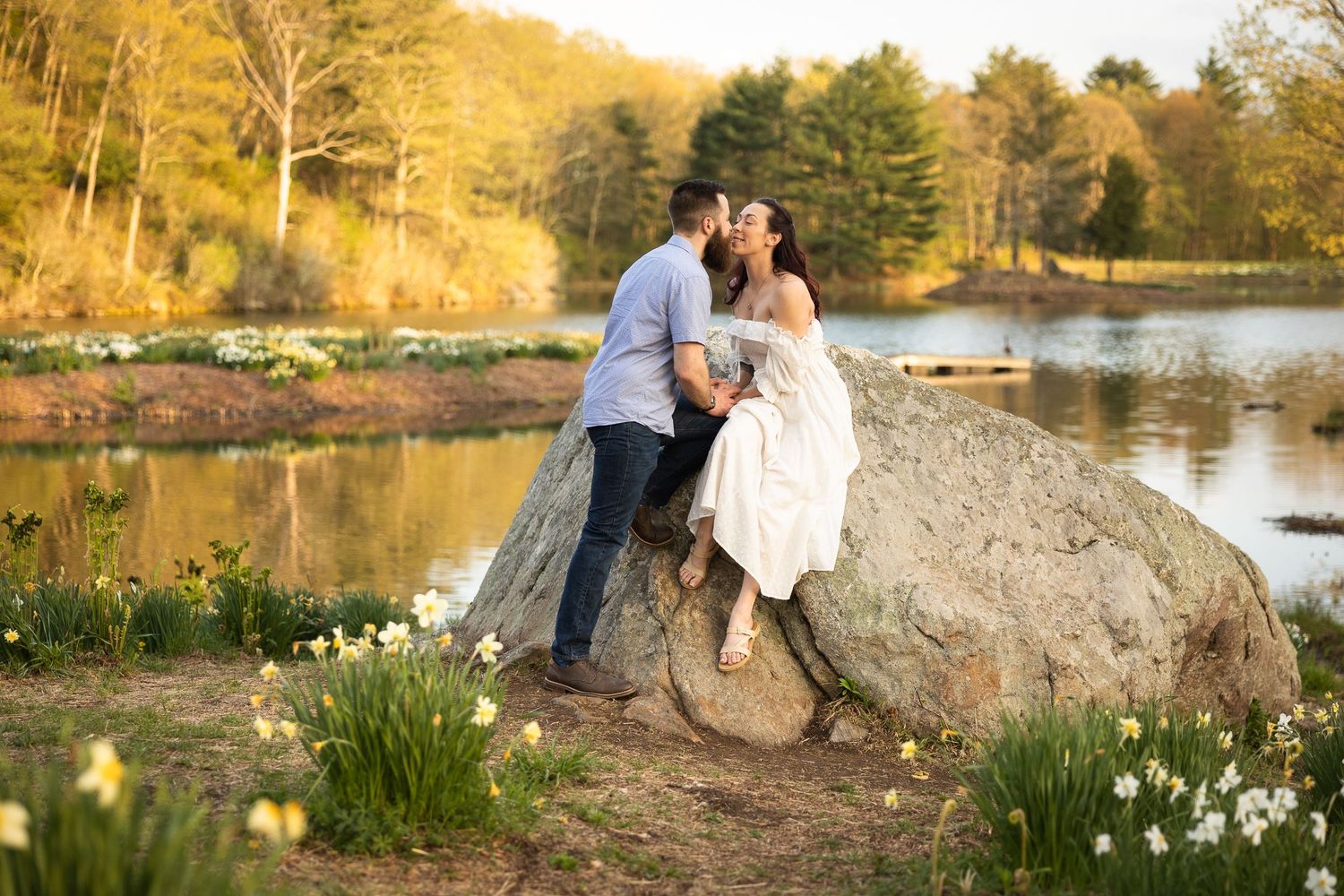 A romantic couple embraces during a golden hour photoshoot by a serene lake with autumn trees in the background.
