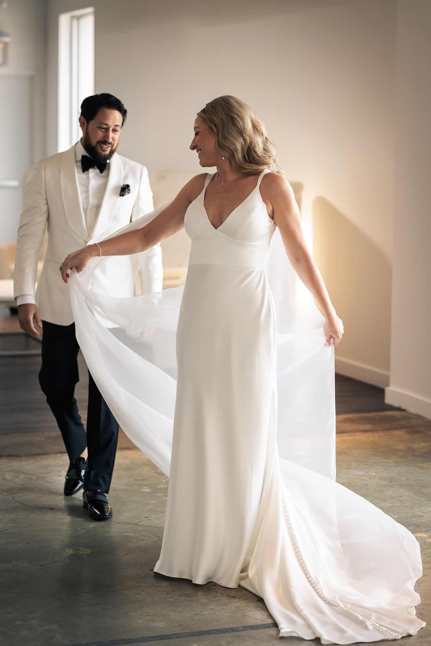 A couple in wedding attire dance together in a sunlit room with white walls and concrete floors.