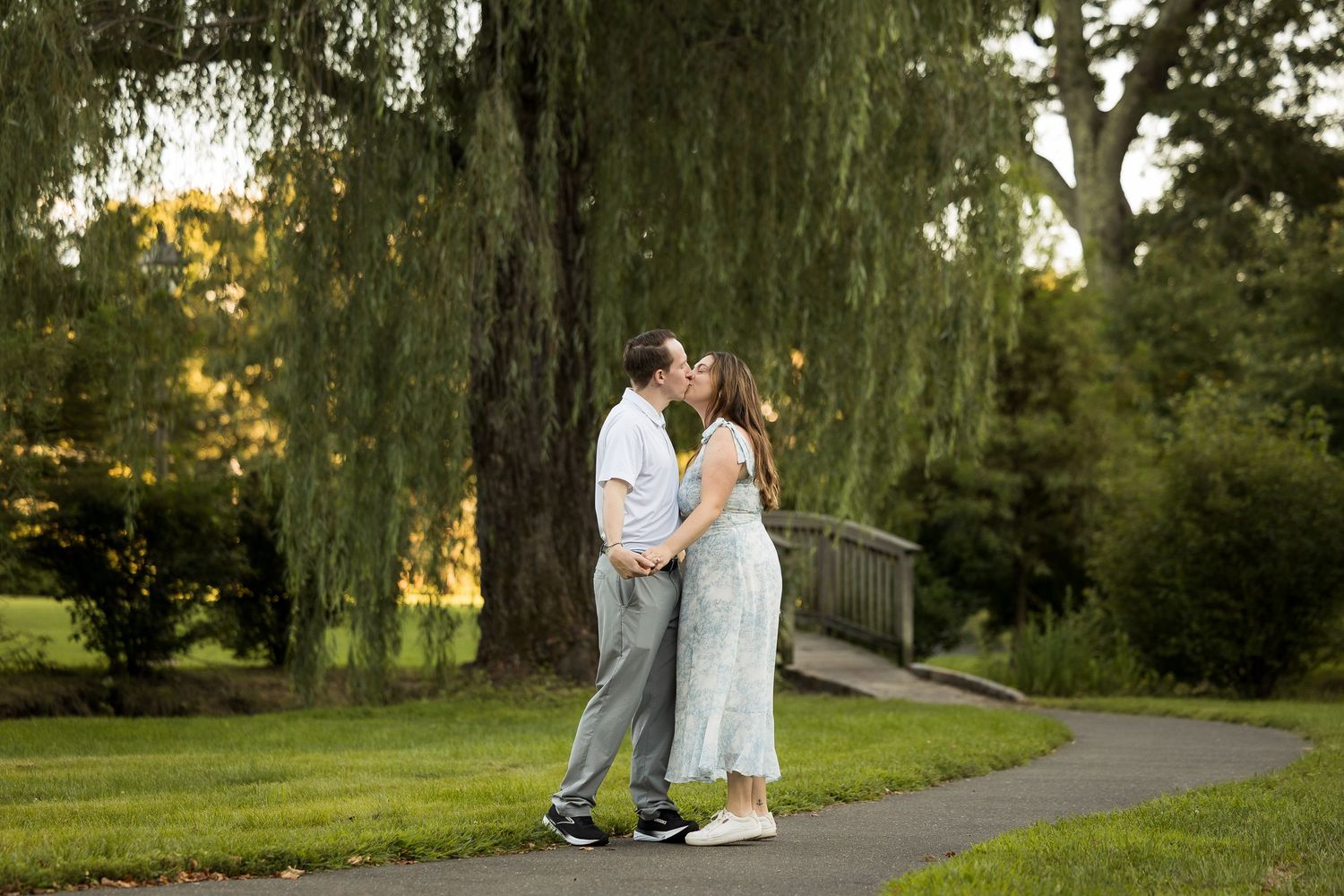 Couple sharing a romantic kiss on a park path under a weeping willow tree at golden hour.