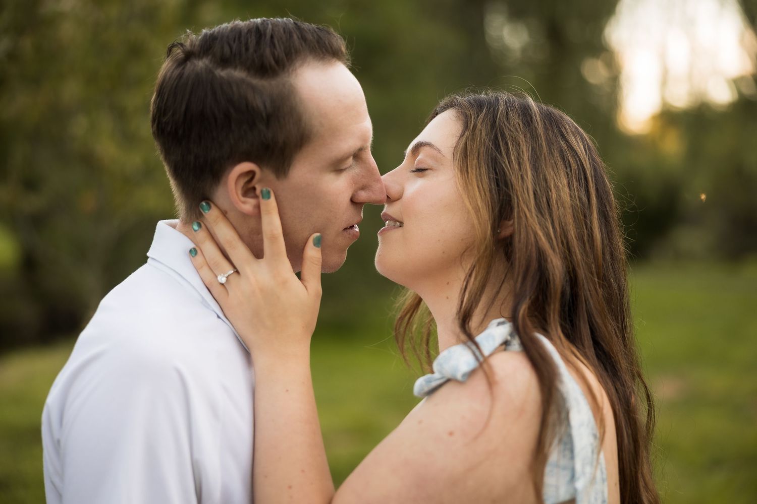 A couple shares a romantic almost-kiss outdoors, with the woman tenderly holding the man's face in a lush green setting.