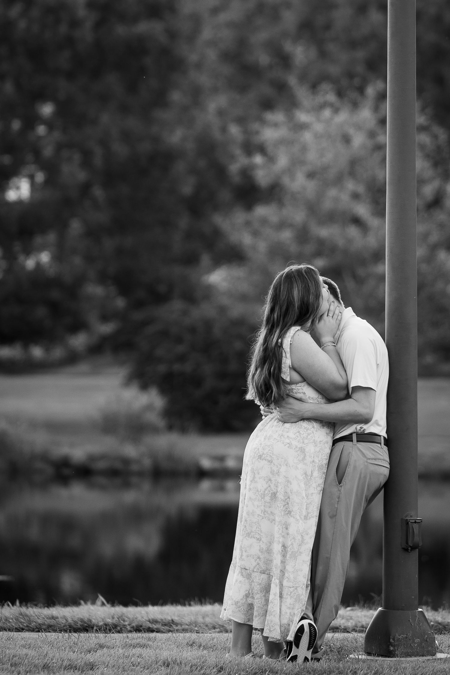 Black and white photo of a couple embracing and kissing near a pole in a lush park setting.