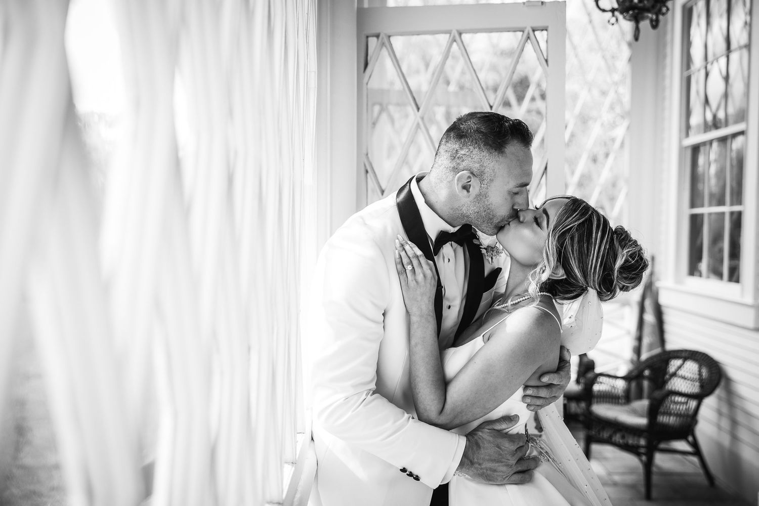 Black and white photograph of a couple sharing an intimate moment on a white porch.