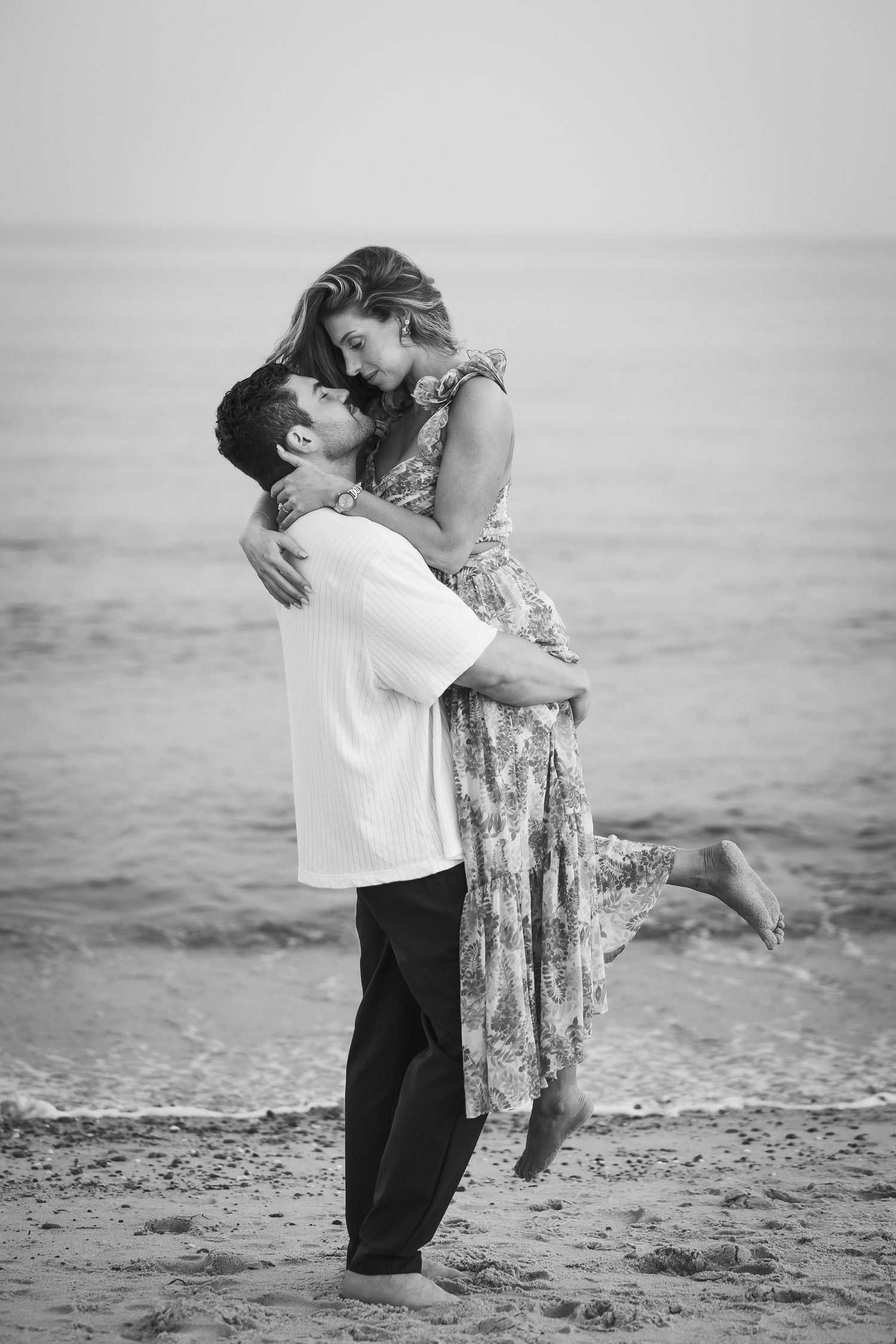 A romantic black and white photo of a couple embracing on the beach, with one person lifted in a floral dress.