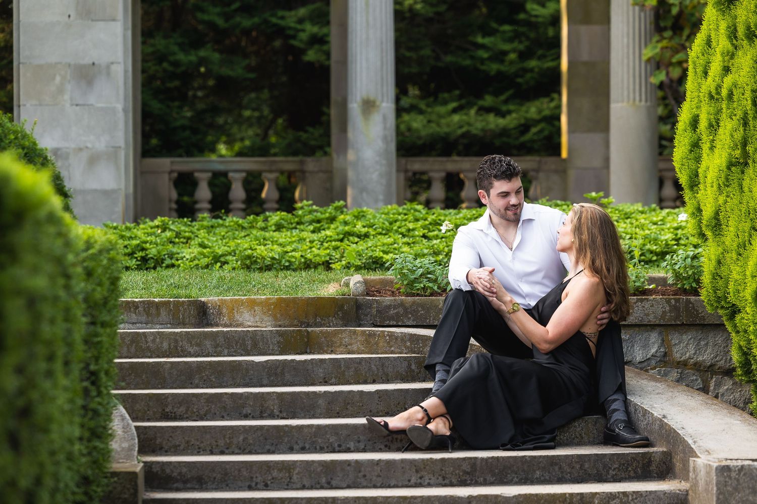Romantic couple sits together on stone steps in a lush garden setting with ornate architectural details.