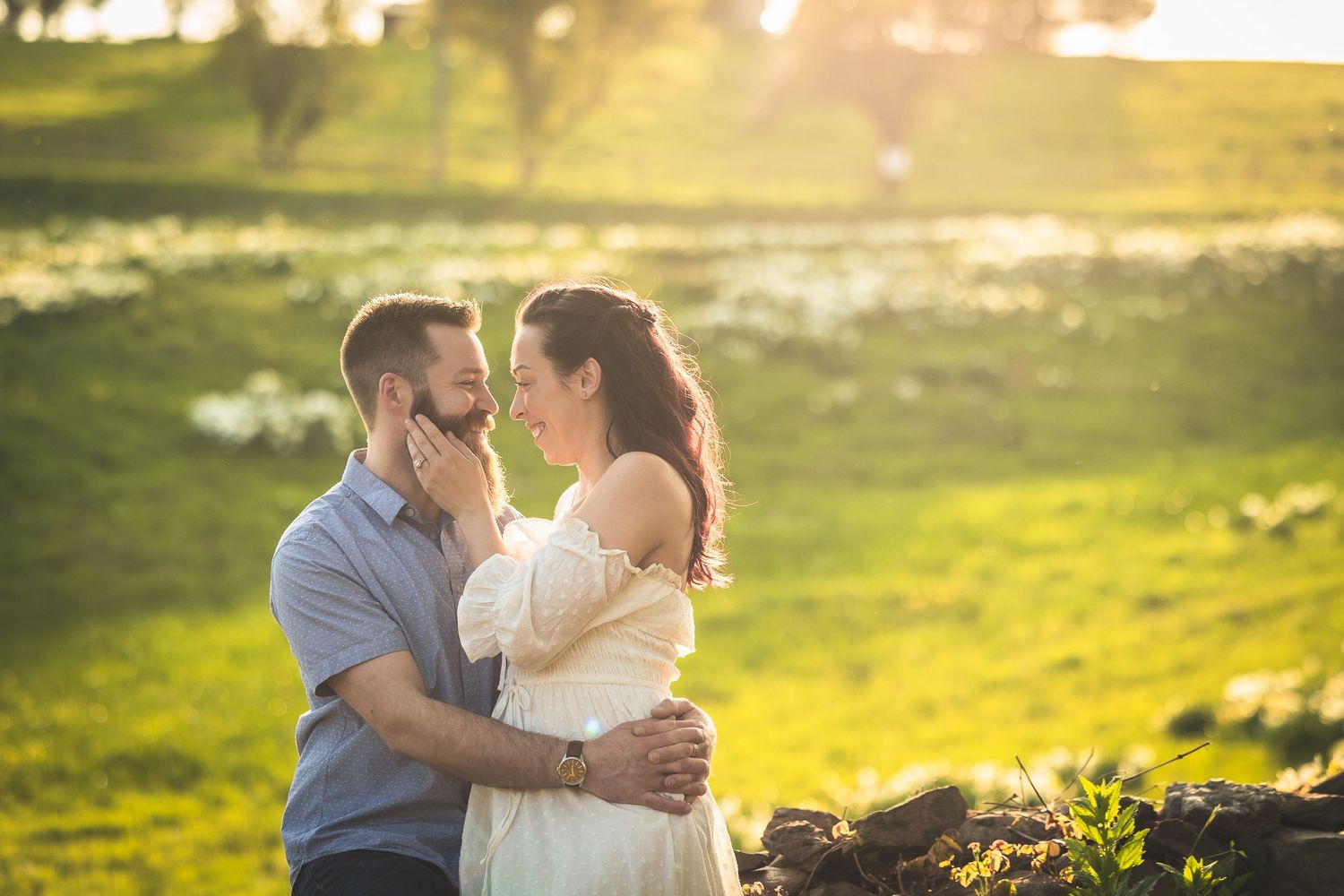 A couple embraces in a sunlit meadow surrounded by white wildflowers during golden hour.