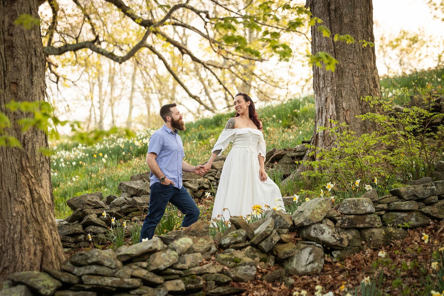 A couple sits together on a rustic stone wall surrounded by autumn trees at sunset.