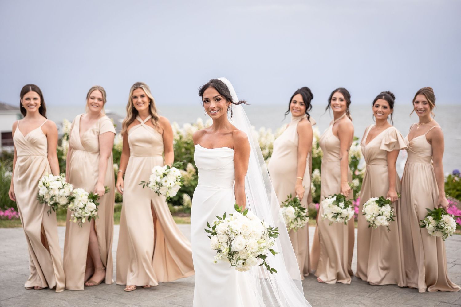Bridal party in champagne colored dresses posing with white bouquets outdoors.