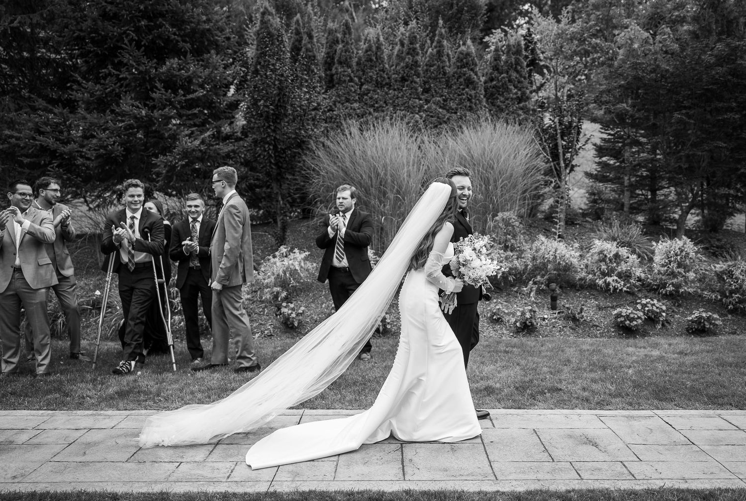 Wedding party and guests gathered for outdoor photos in garden setting.