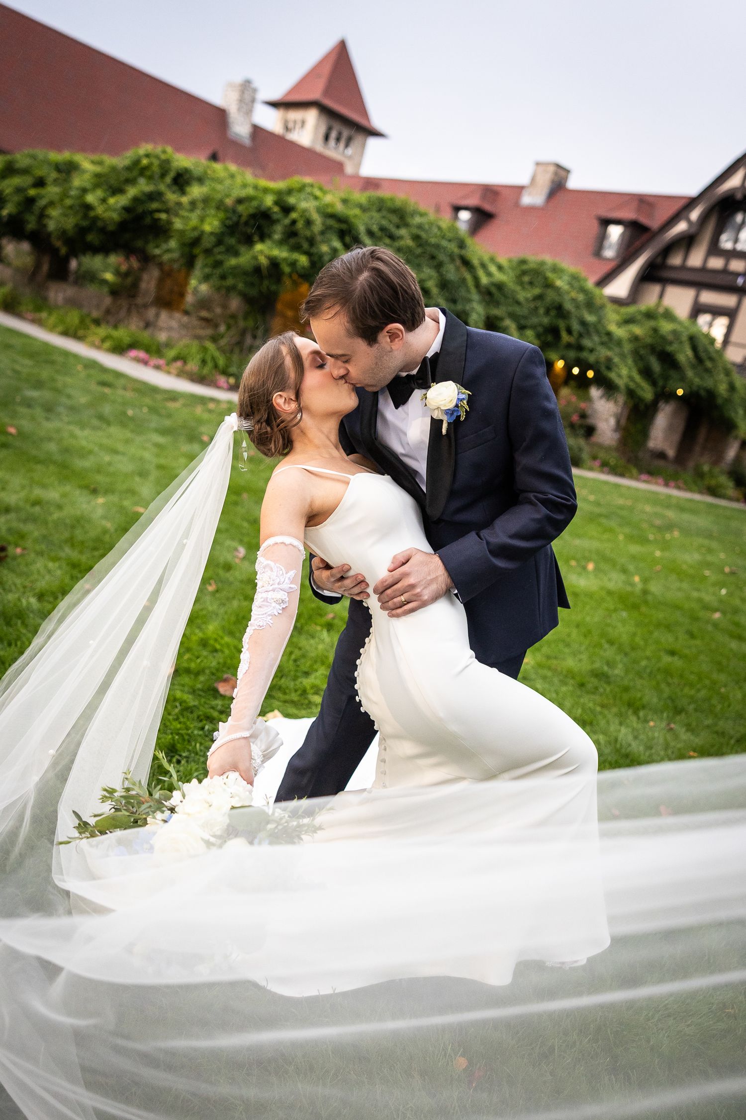 A romantic wedding couple embraces on a green lawn with a dramatic flowing veil creating an elegant scene.