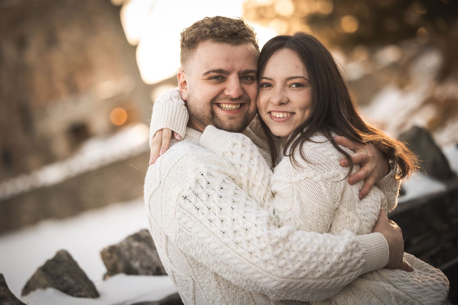 A couple in winter attire embraces during a snowy outdoor photoshoot with warm lighting and bokeh effects.