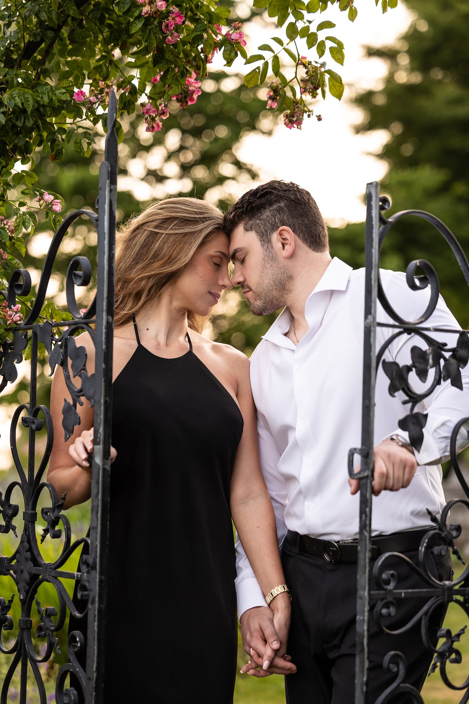 A romantic couple shares an intimate moment by a decorative wrought iron gate in a garden setting.