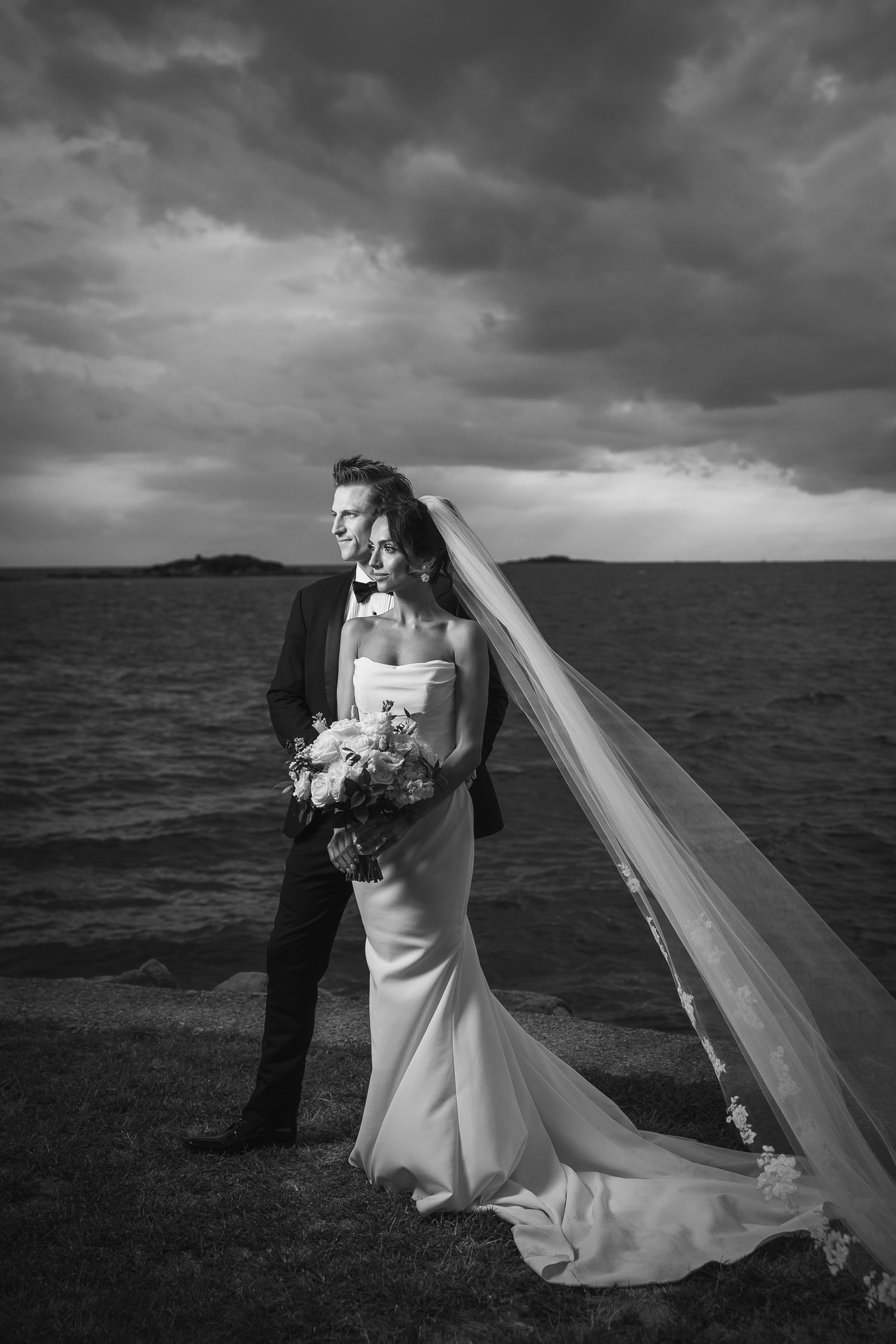 Dramatic black and white coastal portrait with stormy sky and flowing veil.