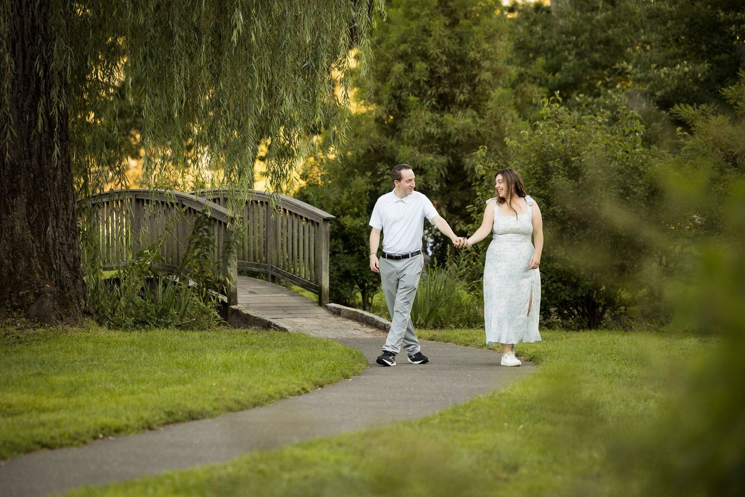 Couple holding hands walking on a garden path near a wooden bridge surrounded by lush green trees.
