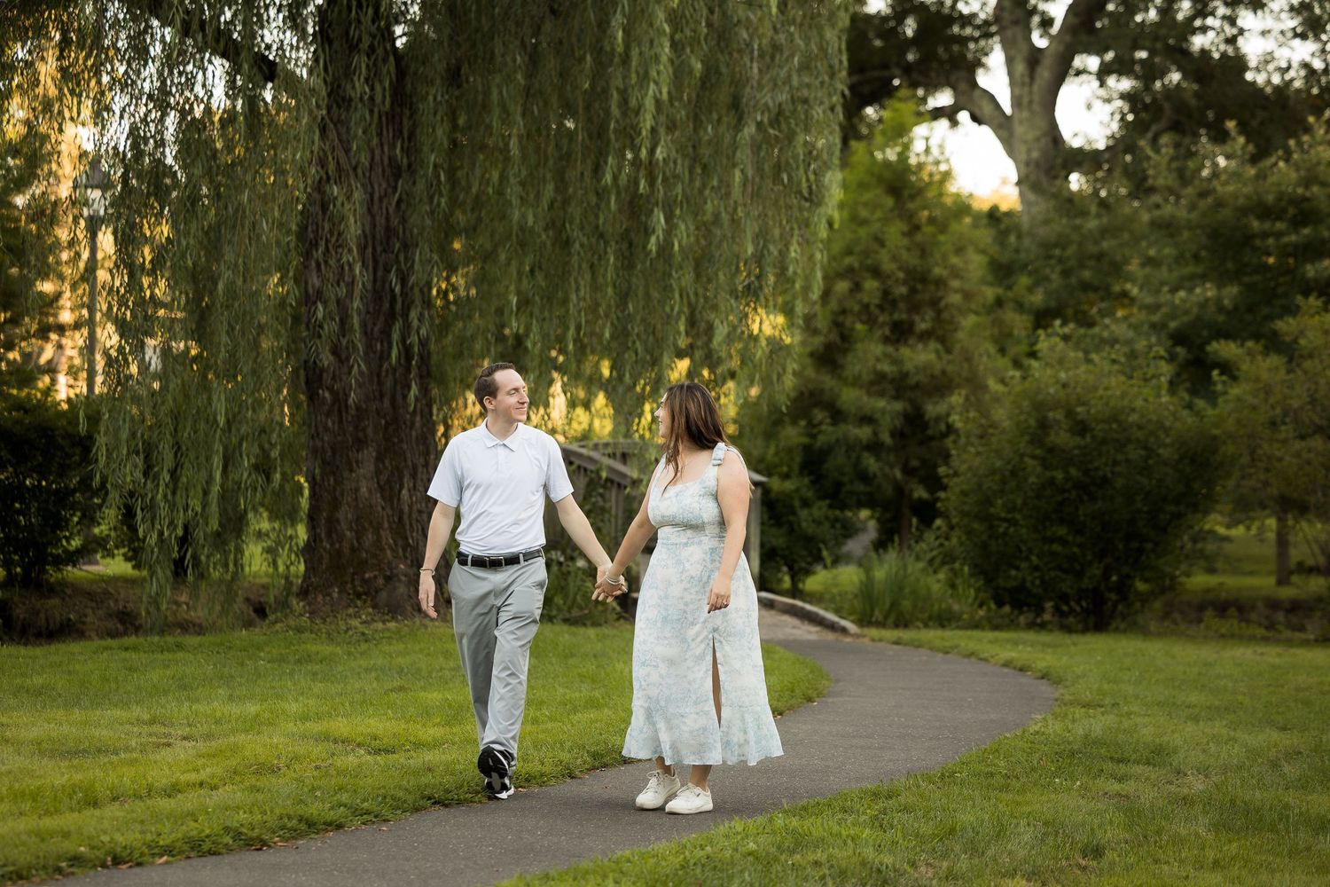Couple holding hands walking on a garden path surrounded by lush green trees and grass during golden hour.