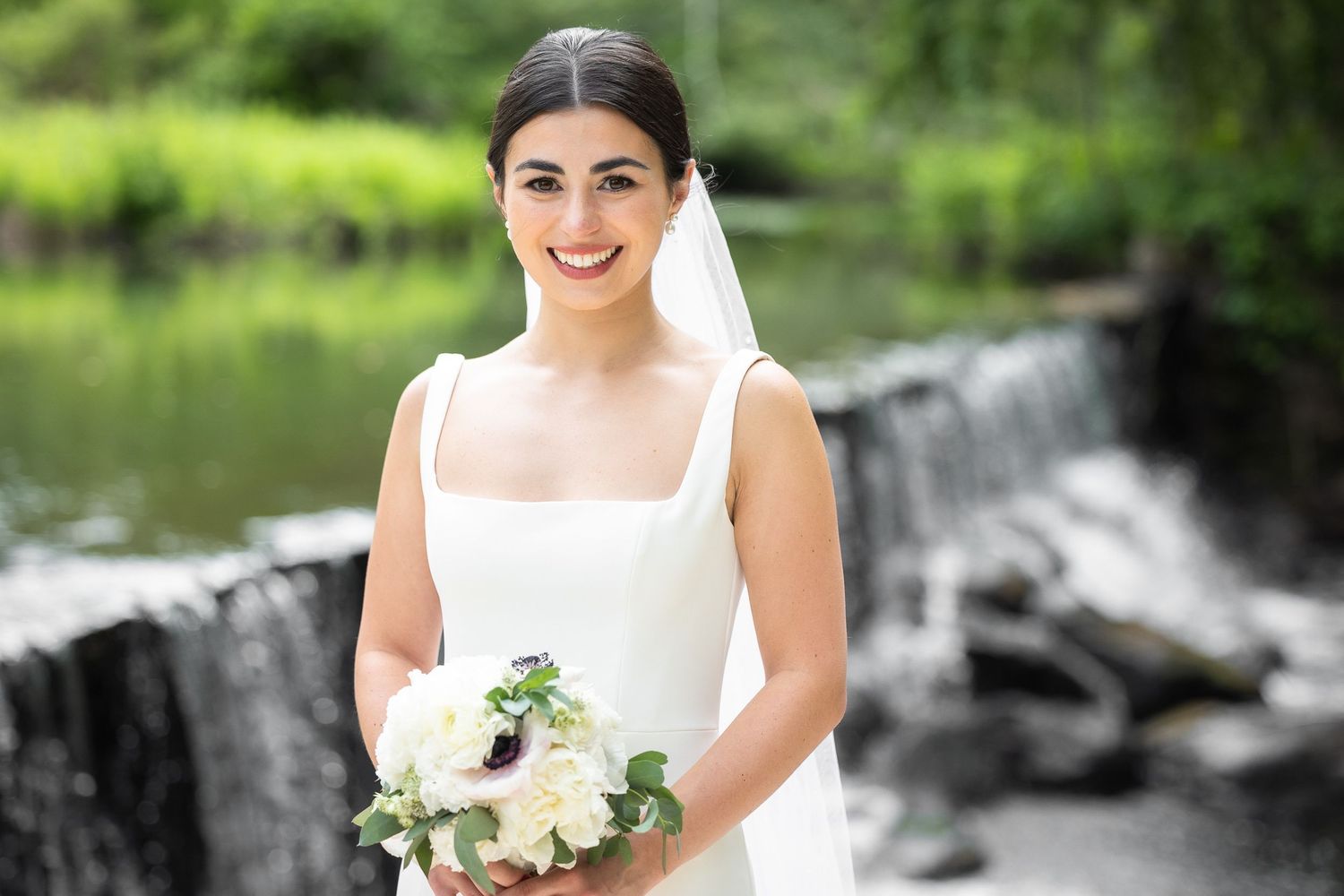 Bride in classic white dress posing near a waterfall in a garden setting.