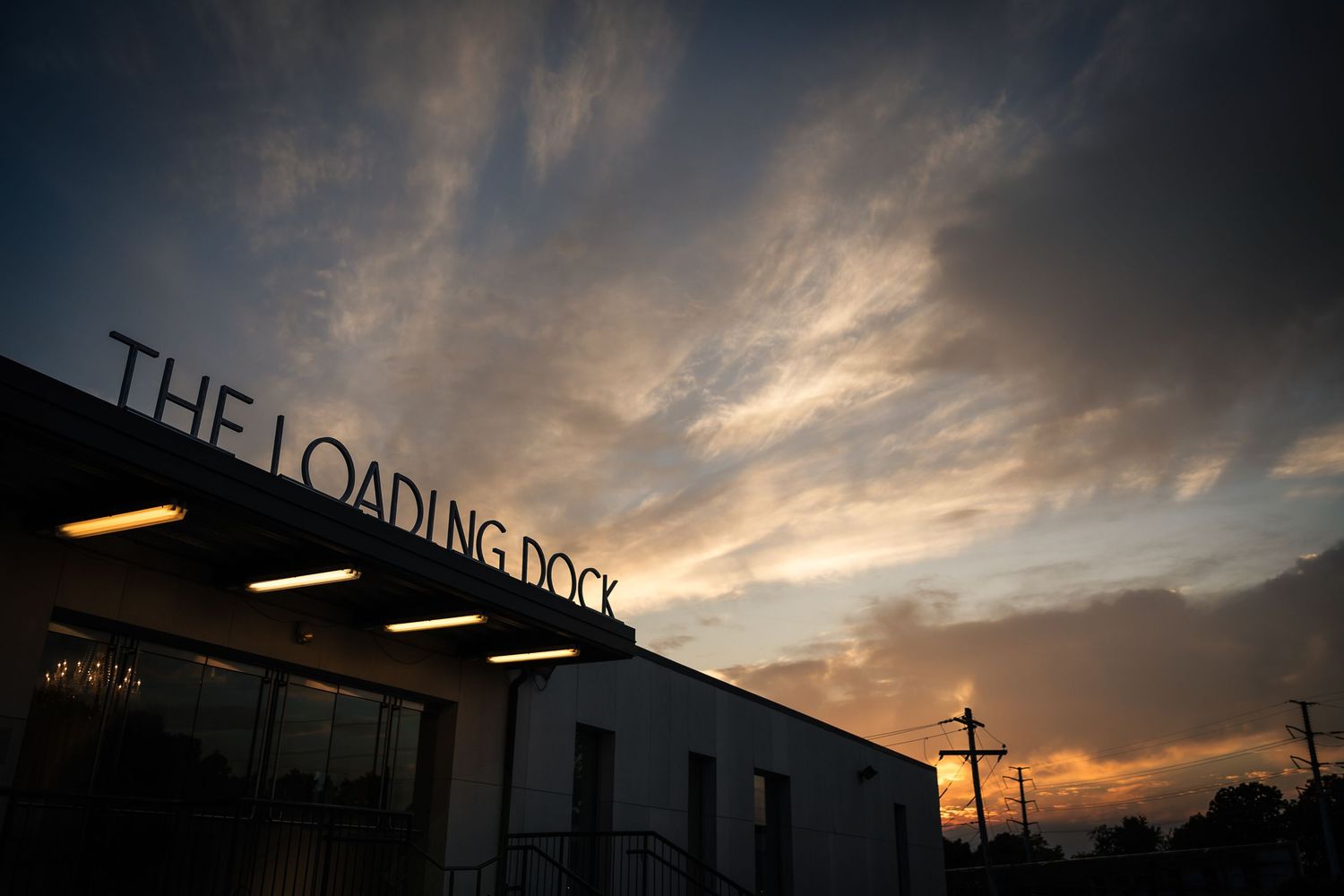 A dramatic sunset sky with wispy clouds above a building with 'THE LOADING DOCK' signage silhouetted against the dusk.
