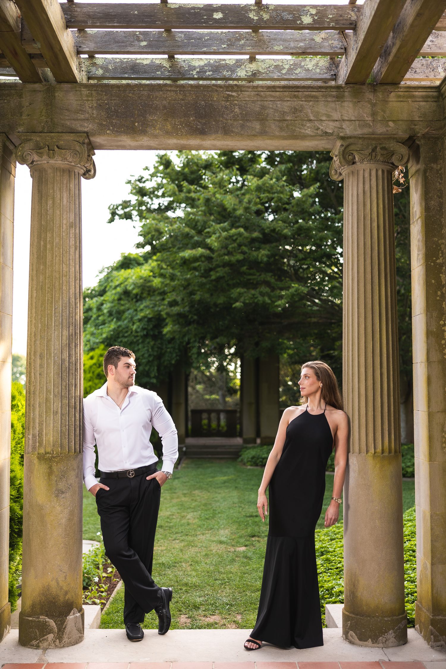 A couple stands between elegant stone columns in a garden pergola, with lush greenery visible in the background.